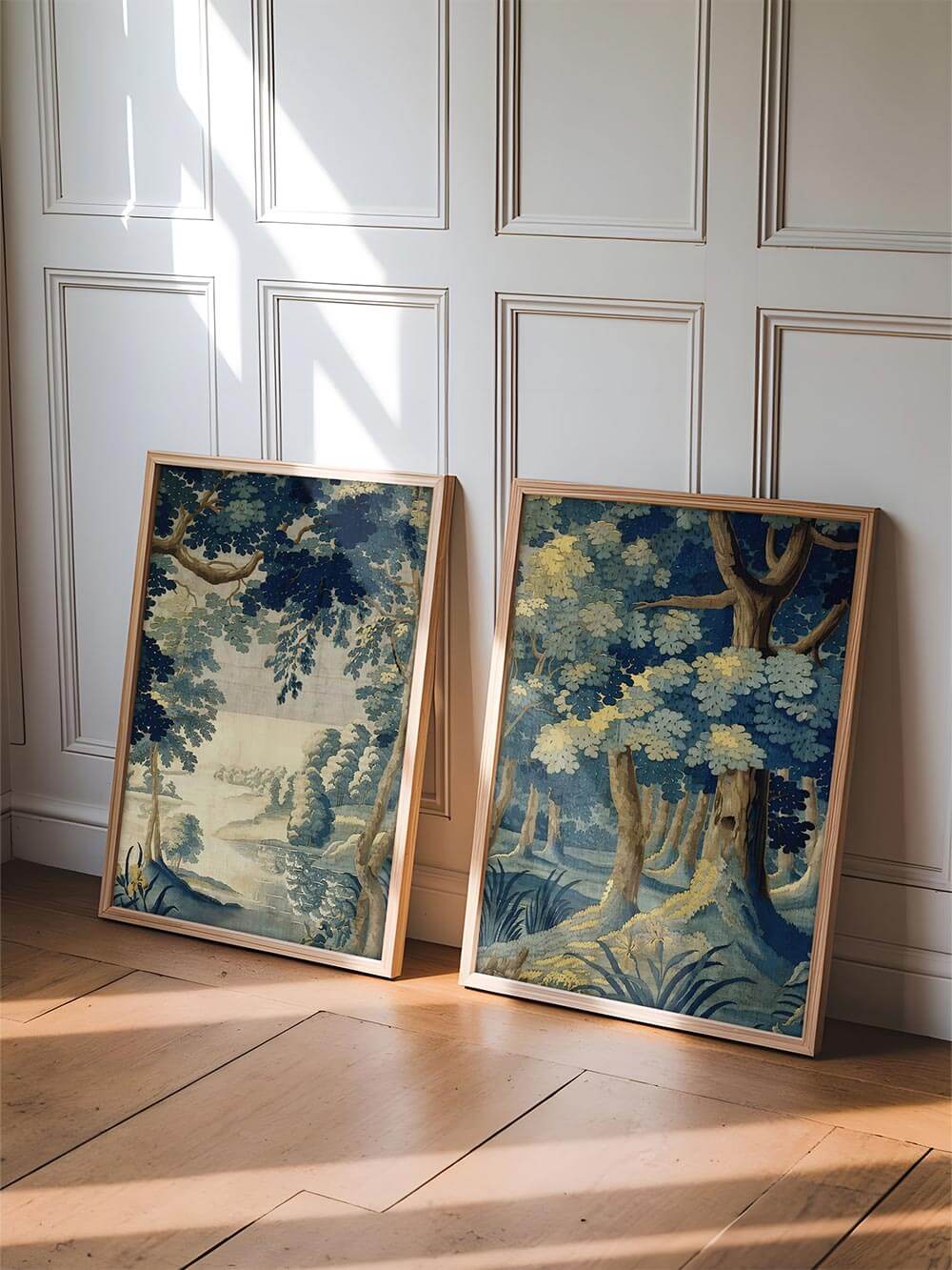 Zwei gerahmte Landschafts-Kunstdrucke mit gold-blauen Waldfarben lehnen im Licht an einer weißen Wandvertäfelung auf Holzboden.