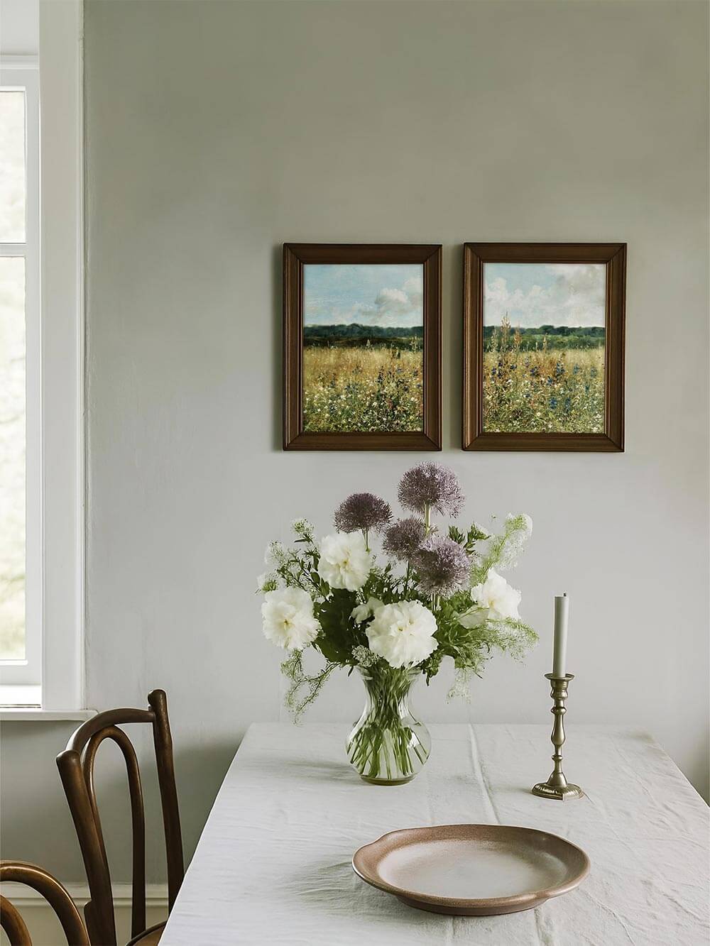 Zwei Vintage-Kunstdrucke mit Wiesenblumen hängen über einem gedeckten Esstisch mit Blumenstrauß. Die Motive ergänzen den ruhigen Landhausstil.