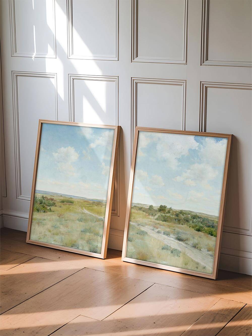 Zwei gerahmte Landschafts-Poster lehnen an einer Wand im Sonnenlicht. Die hellen Holztöne und die naturinspirierte Szene strahlen Leichtigkeit und ländliche Eleganz im Wabi-Sabi-Stil aus.