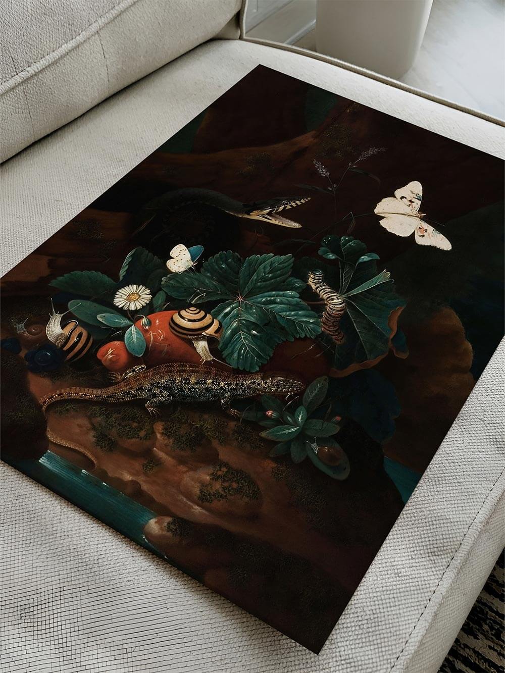 Kunstdruck mit dunkler botanischer Szene liegt ungerahmt auf einem hellen Sofa. Die Komposition aus Schmetterlingen, Schnecken und Blättern wirkt lebendig und nostalgisch.