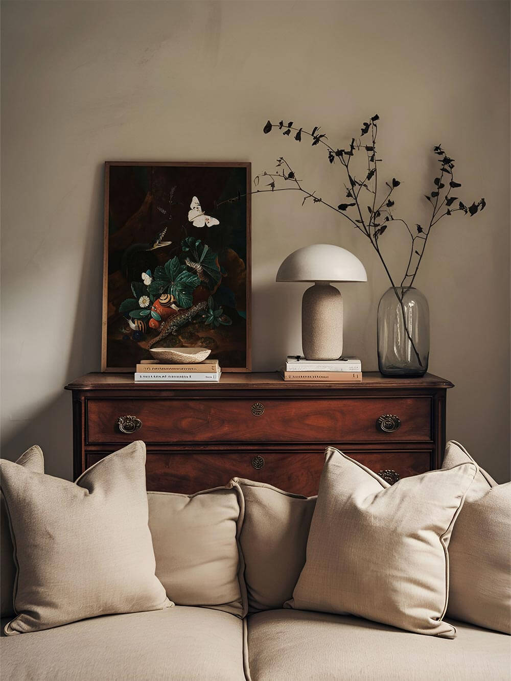 Der dunkle botanische Kunstdruck steht auf einer antiken Kommode vor einer beigen Wand. Daneben setzen ein Zweig in Glasvase und eine Pilzlampe ruhige Akzente im Raum.
