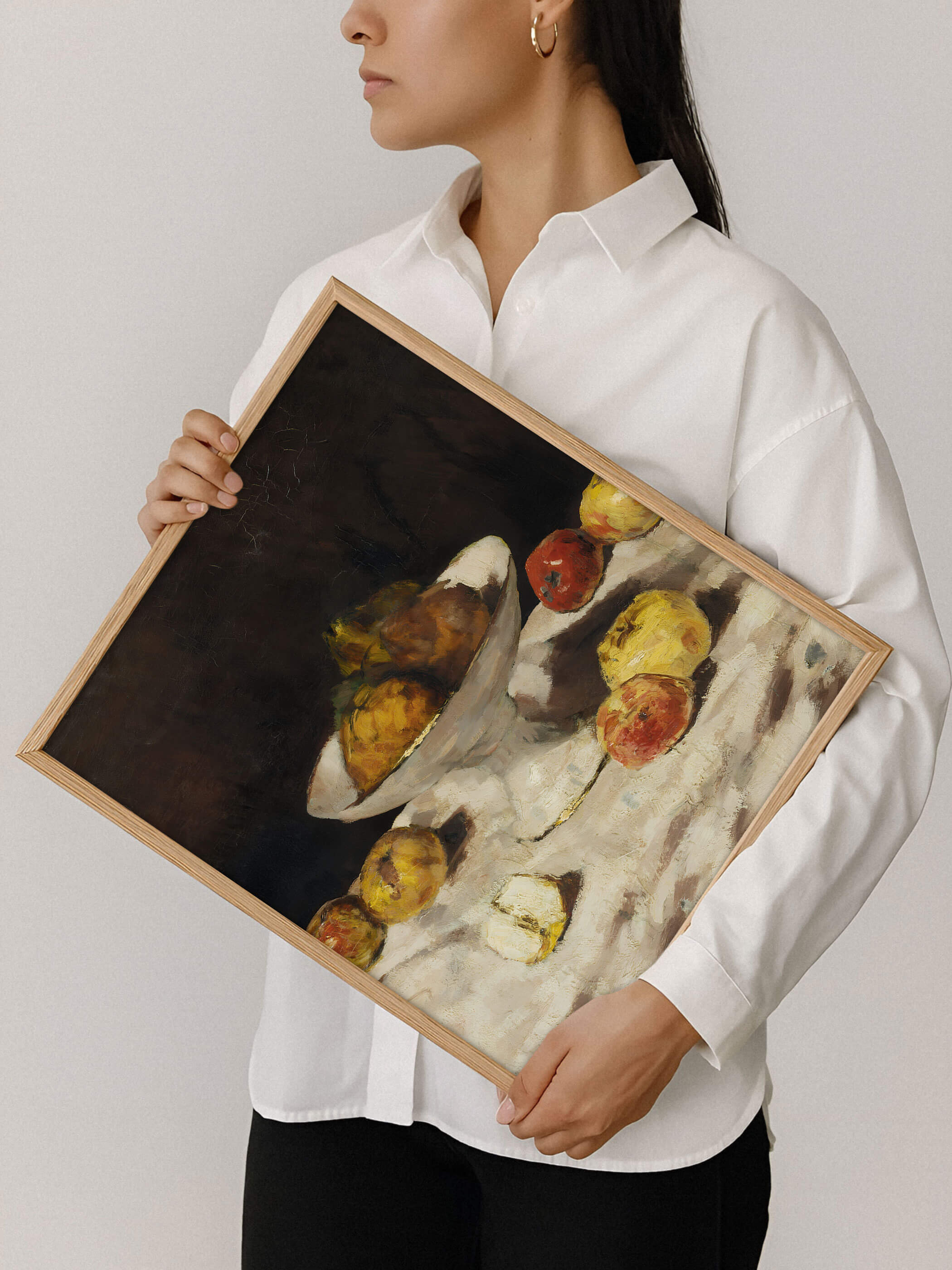 Eine Frau in weißem Hemd hält einen Holzrahmen mit dem Stillleben aus Äpfeln, Birnen und einer weißen Kompottschale vor dunklem Hintergrund. Der Kunstdruck zeigt die naturbelassene Holzrahmung und die satten, warmen Farbtöne des Motivs im Detail.