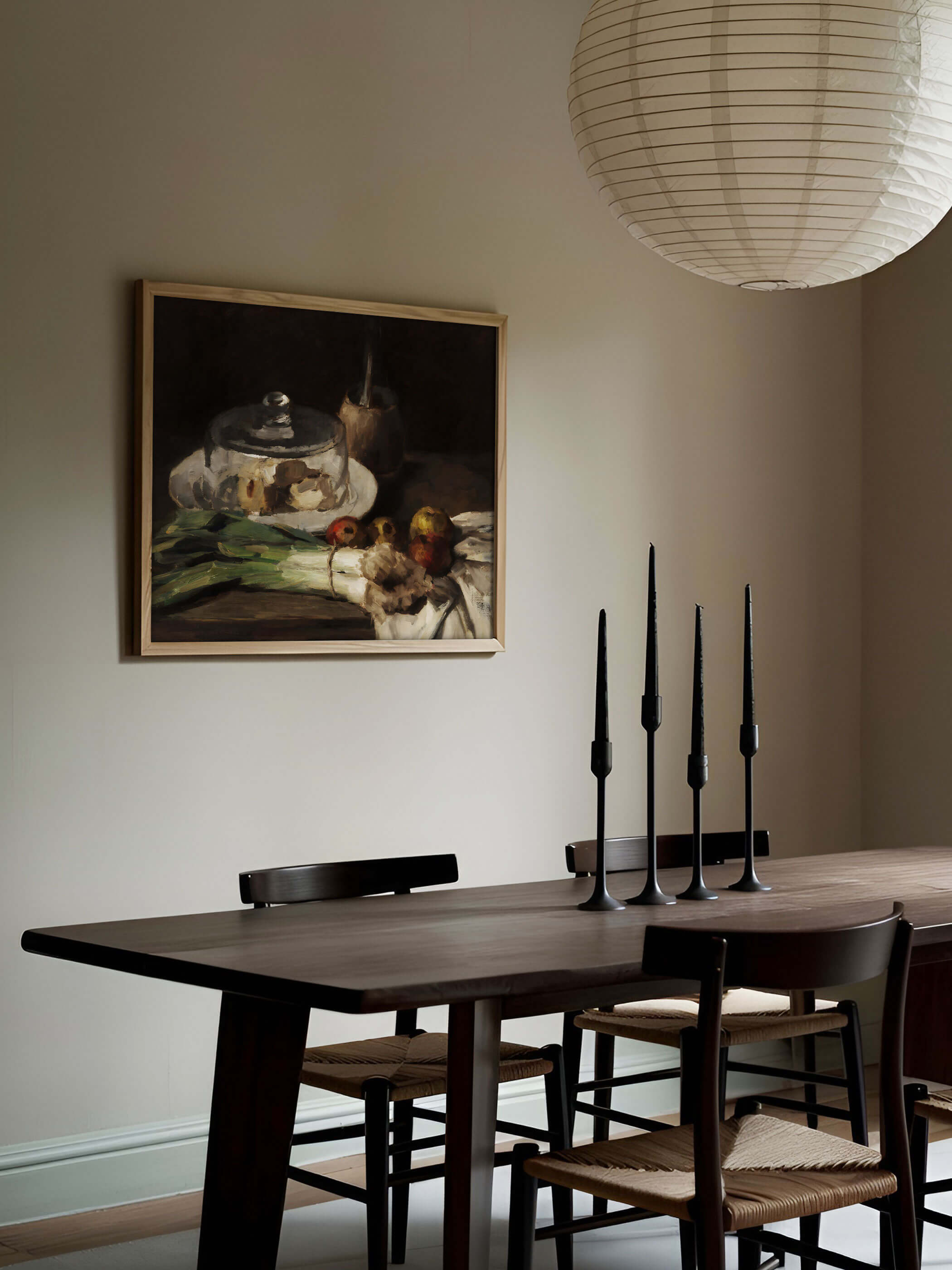 Hell gerahmter Kunstdruck mit Küchenstillleben hängt an einer beigen Wand über einem dunklen Holztisch mit schwarzen Kerzenständern und Papierlampen. Das ruhige Vintage Wandbild fügt sich in die reduzierte, warme Esszimmergestaltung ein.