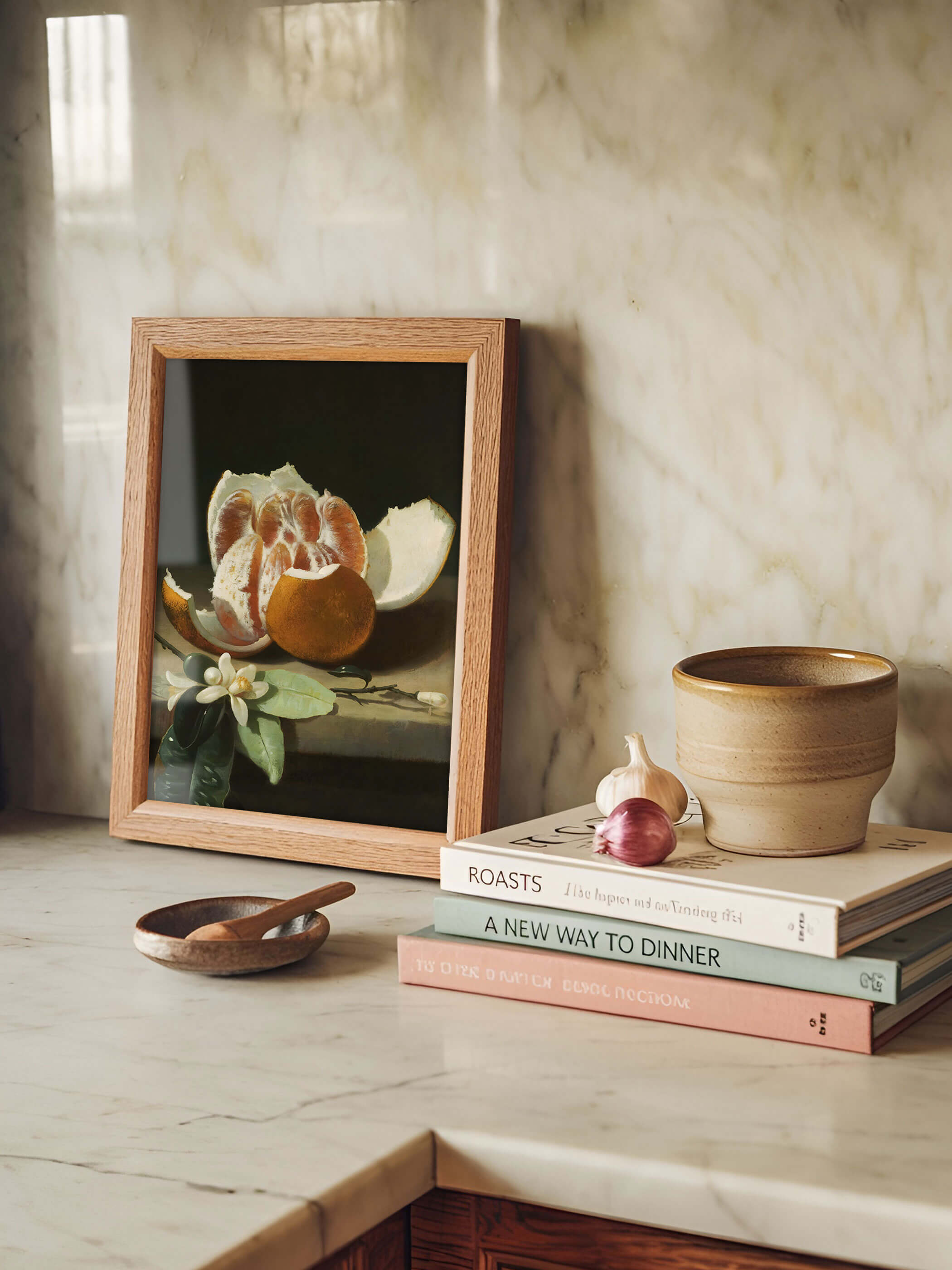 Gerahmter Kunstdruck mit geschälter Orange und Orangenblüte lehnt auf einer Marmorarbeitsplatte neben Kochbüchern, Knoblauch und einer Keramikschale. Der warme Holzrahmen und das dunkle Barock-Motiv setzen einen ruhigen Akzent in der hellen Küche.