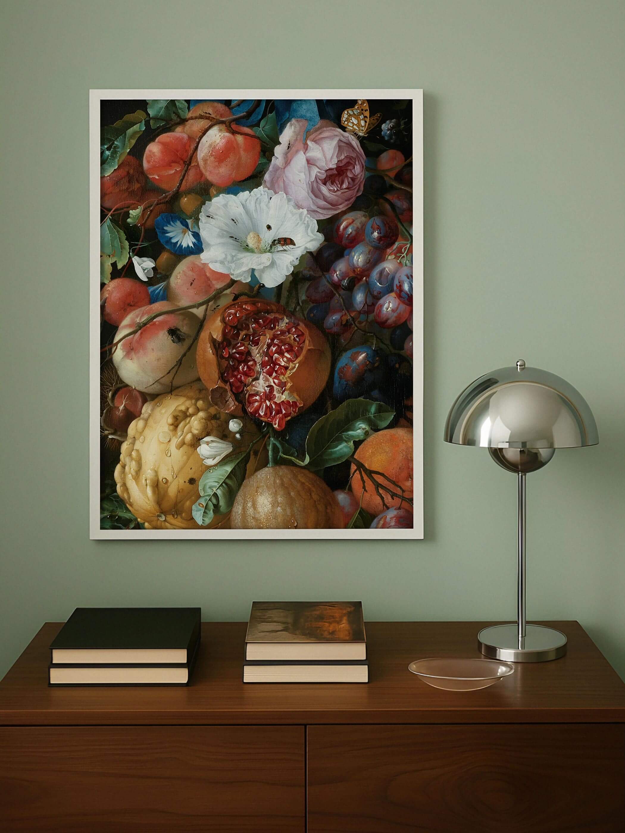 Cremefarben gerahmter Kunstdruck mit barocker Früchtegirlande hängt an einer salbeigrünen Wand über einer dunklen Holzkommode mit Büchern und einer Chromlampe. Die satten Rot- und Blautöne des Motivs bilden einen lebendigen Kontrast zur ruhigen Wandfarbe.