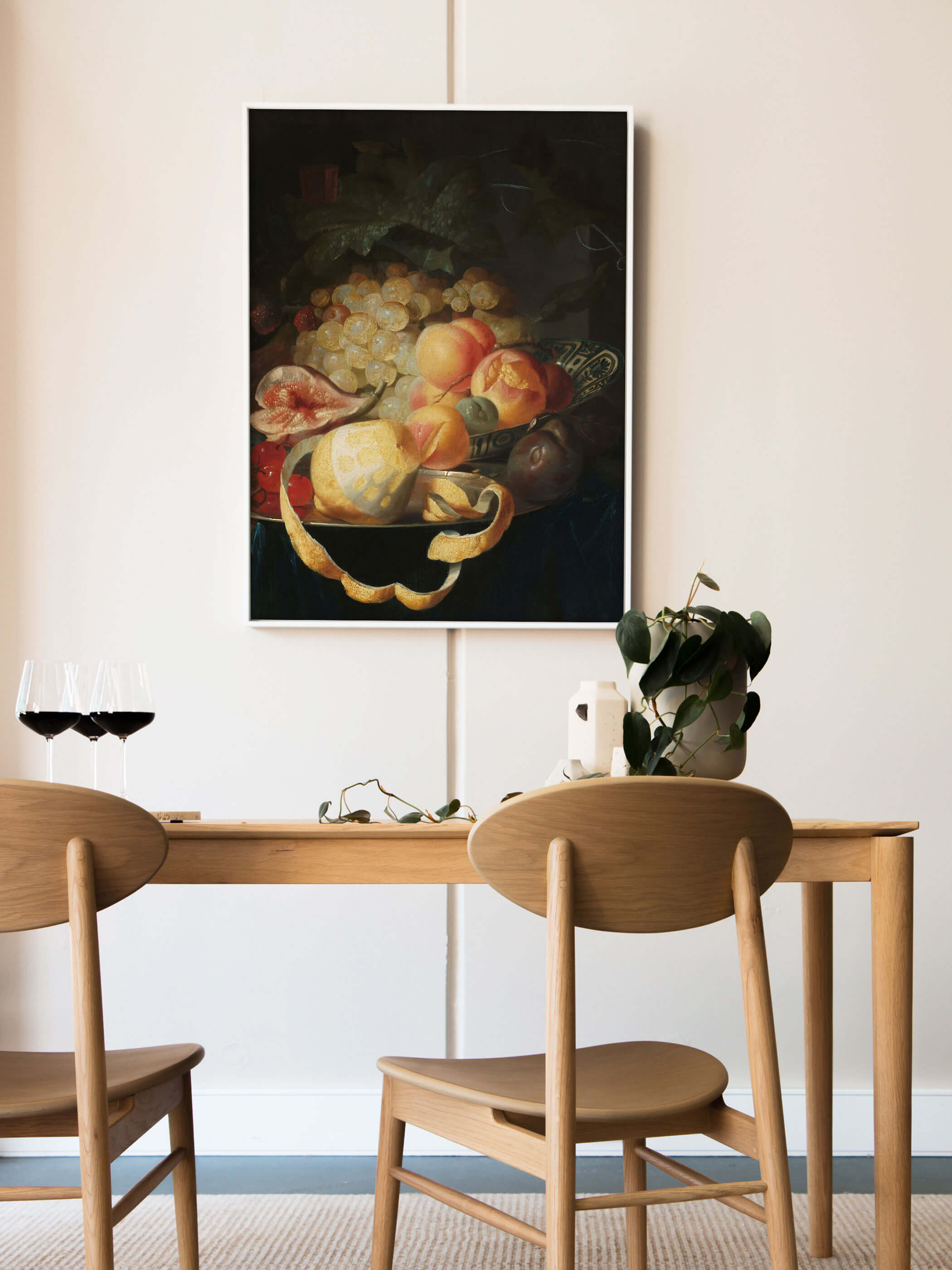 Weiß gerahmter Kunstdruck mit barockem Fruchtstillleben lehnt an der Wand über einem Holztisch mit Rotweingläsern und einer grünen Pflanze. Das dunkle, edle Motiv setzt einen eleganten Akzent im hellen Esszimmer.