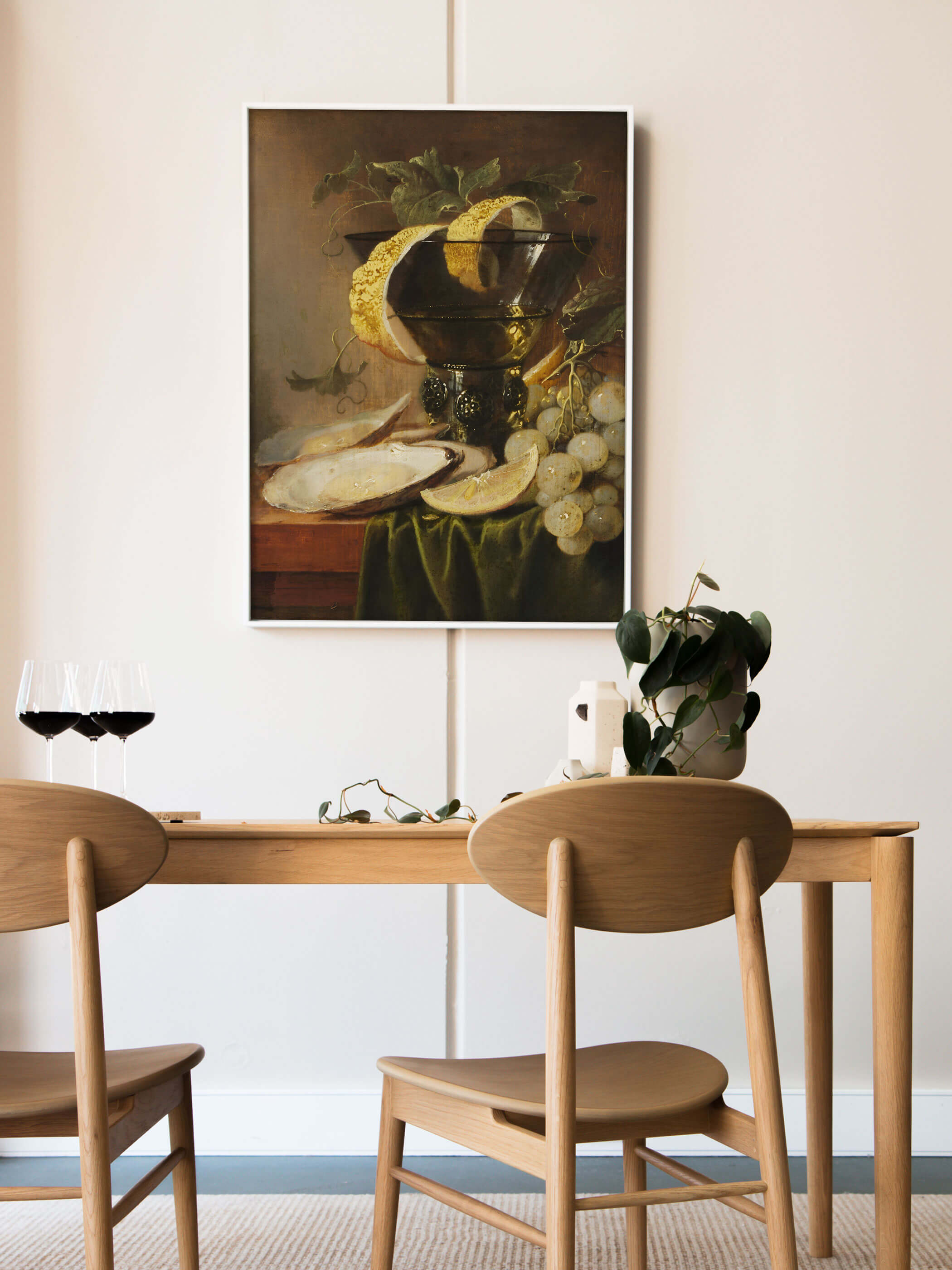 Weiß gerahmter Kunstdruck mit barockem Austern-Stillleben lehnt an der Wand über einem Holztisch mit Rotweingläsern und einer grünen Pflanze. Das dunkle, edle Motiv setzt einen stilvollen Akzent im hellen Esszimmer.