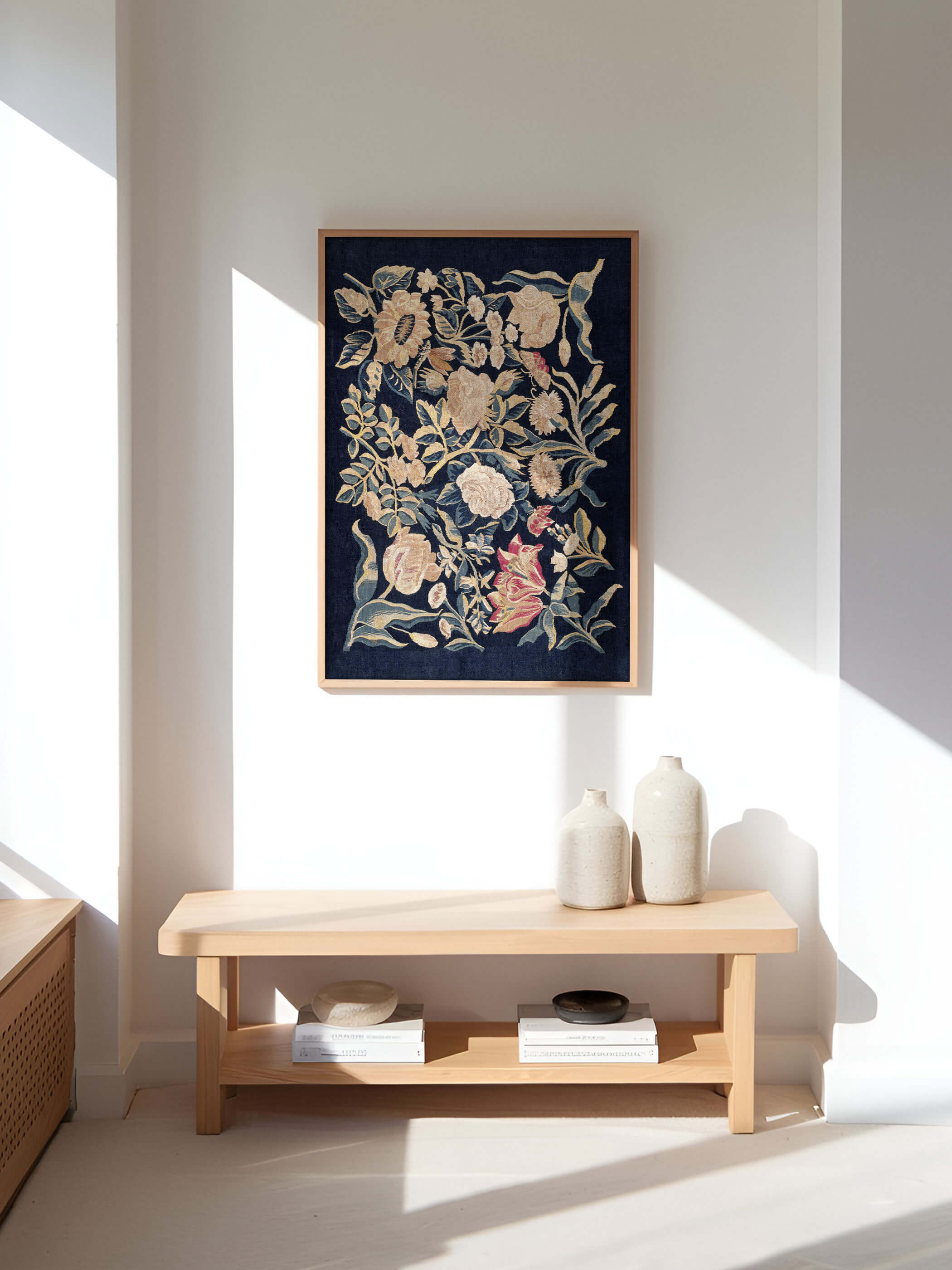 Vintage-Textilposter mit Blumenmotiven an einer hellen weißen Wand, eingerahmt in Holz. Die Blumen in Beige und Rosa heben sich von einem dunklen Hintergrund ab. Minimalistische Dekoration mit Keramikvasen auf einem Holzregal darunter.