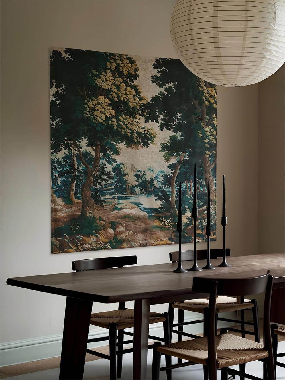 Eleganter Verdure-Wandteppich in einem dunklen, stimmungsvollen Esszimmer mit schwarzen Kerzenhaltern und Holztisch. Das üppige Landschaftsmotiv mit Bäumen und Fluss fügt sich perfekt in ein luxuriöses, klassisches Interieur.