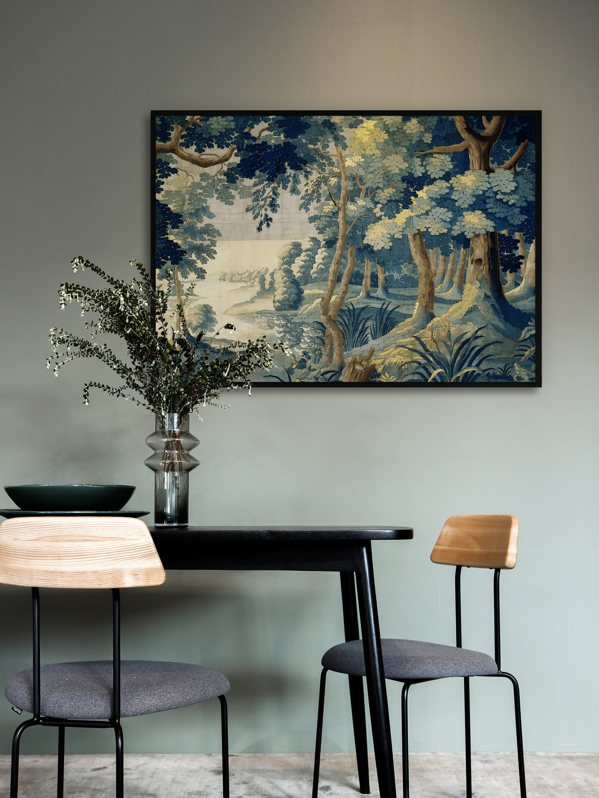 Großes Poster mit blauer Verdure Landschaft im schwarzen Rahmen an einer salbeigrünen Wand über einem schwarzen Esstisch mit zwei Stühlen aus hellem Holz. Eine Glasvase mit grünen Zweigen nimmt das Grün des Motivs auf.
