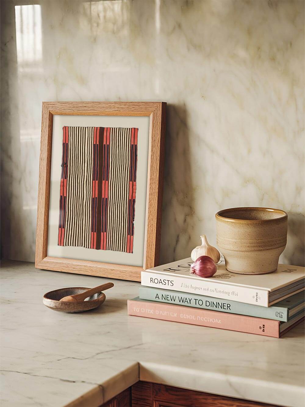 Gerahmter Kunstdruck mit grafischem Streifenmuster steht auf einer Marmorarbeitsfläche neben Keramik und Kochbüchern. Das textile Wandbild ergänzt die Küche mit warmen Farben und einer ruhigen, handwerklich inspirierten Atmosphäre.