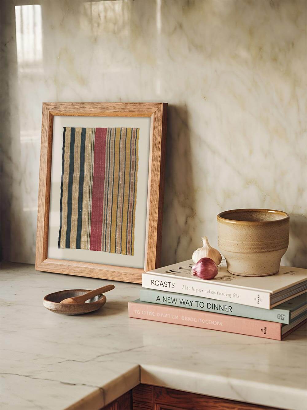Textiler Kunstdruck mit handgewebter Streifenstruktur lehnt auf einer Marmorarbeitsfläche neben Keramik und Kochbüchern. Die erdigen Farben und die sichtbare Webstruktur verleihen dem Raum eine ruhige, authentische Wohnlichkeit.