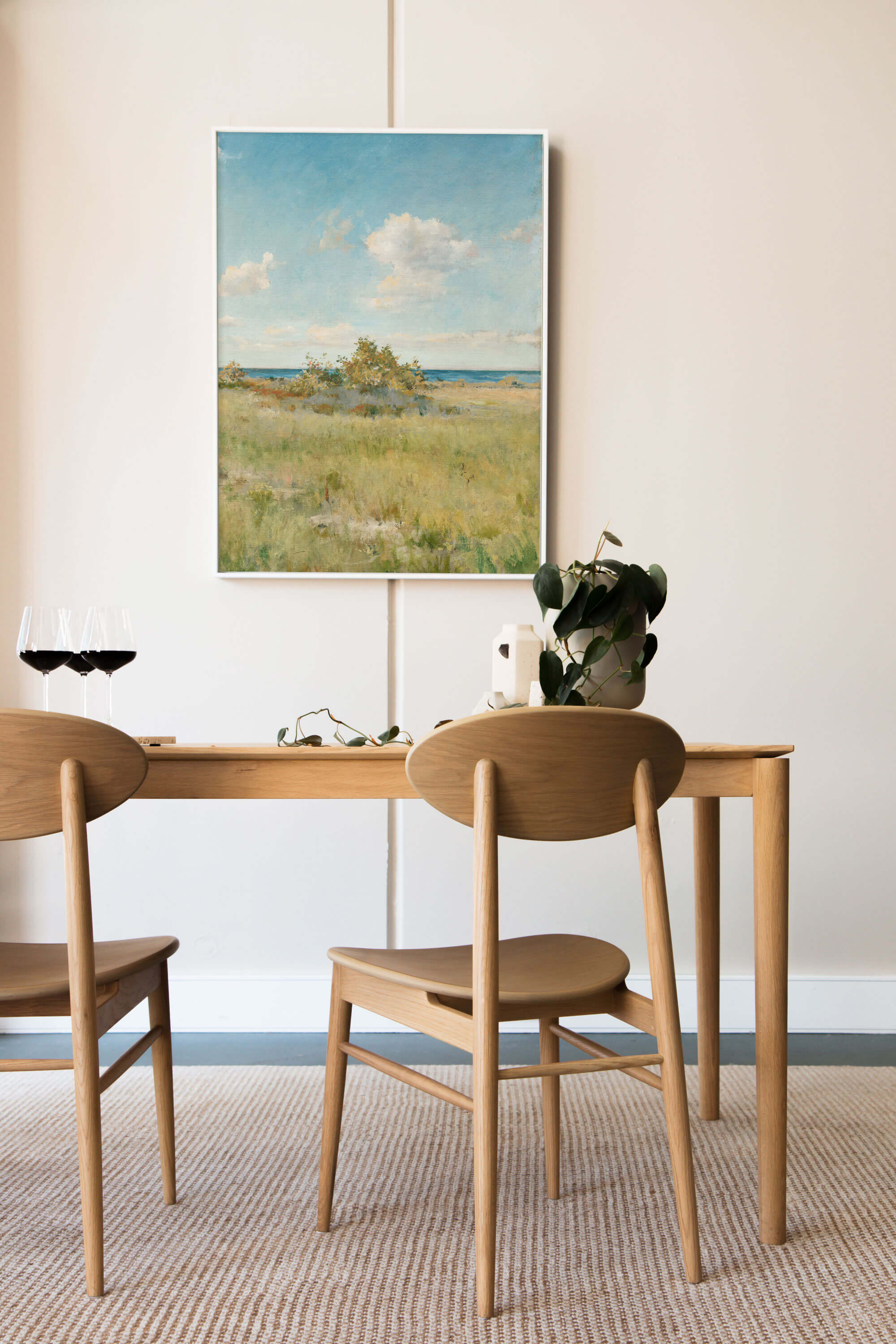 Weiß gerahmter Kunstdruck mit sonniger Küstenlandschaft hängt an einer cremefarbenen Wand über einem Holztisch mit Rotweingläsern und einer grünen Pflanze. Das helle, luftige Motiv setzt einen ruhigen Akzent im Esszimmer.