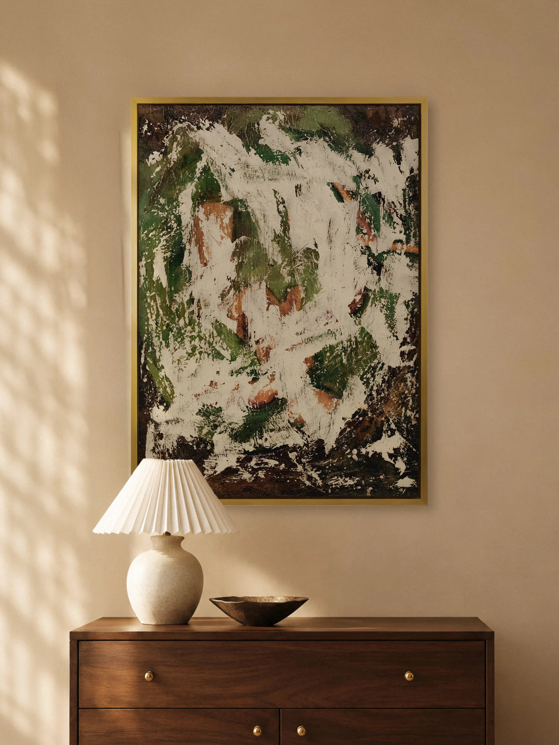 Abstraktes Leinwandbild in Grün und Creme im Goldrahmen über einer Kommode aus dunklem Holz mit Keramiklampe und Metallschale. Das Wabi Sabi Wandbild wirkt im warmen Sonnenlicht erdig und ruhig.
