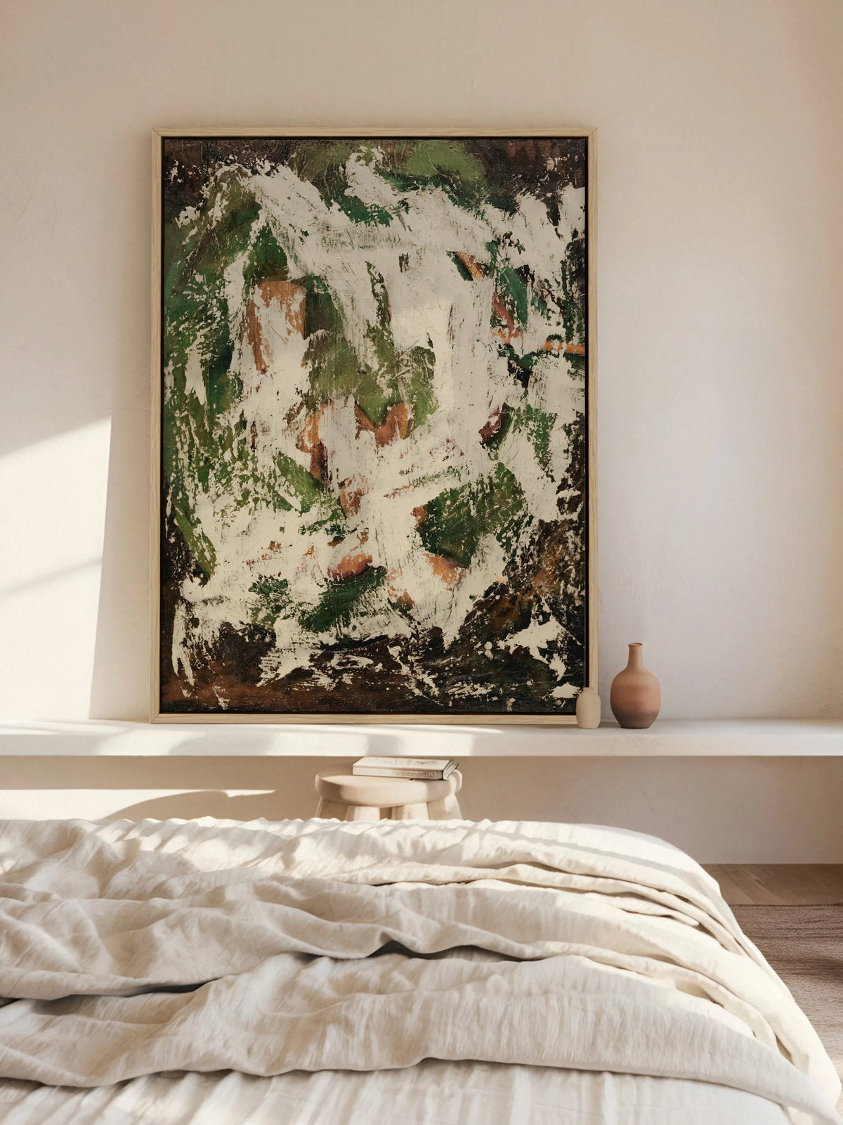 Abstraktes Leinwandbild im Naturholzrahmen lehnt auf einer weißen Wandablage über einem Bett mit Leinenbettwäsche. Das grüne Wabi Sabi Wandbild bringt Farbe in das helle Schlafzimmer.