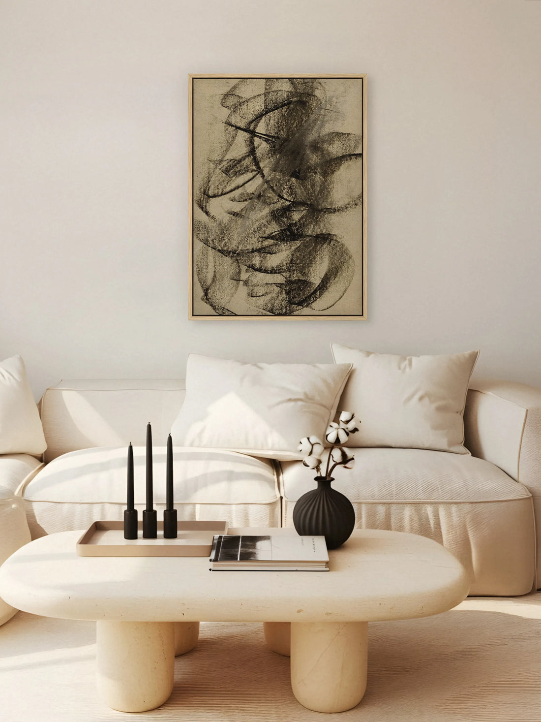Abstraktes Leinwandbild im Naturholzrahmen über einem cremefarbenen Sofa, davor ein ovaler Couchtisch mit schwarzen Kerzen und Baumwollzweigen. Das Wabi Sabi Wandbild greift das Schwarz der Deko auf.