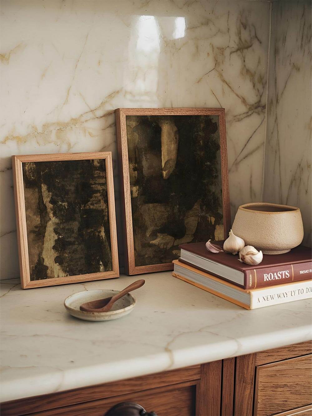Zwei gerahmte abstrakte Kunstdrucke in dunklen Erdtönen lehnen auf einer hellen Marmorarbeitsfläche neben Keramik und Kochbüchern. Die ruhige, natürliche Farbpalette unterstreicht den Wabi-Sabi Stil und fügt sich harmonisch in eine minimalistische Küche ein.