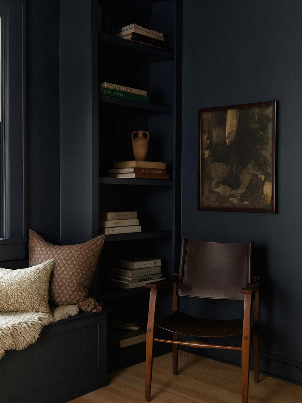 Dunkler abstrakter Kunstdruck mit erdigen Texturen hängt in einem moody Wohnzimmer mit tiefblauen Wänden, Bücherregal und Holzsessel. Das Wandbild schafft eine ruhige, kontemplative Stimmung und passt ideal zu Wabi-Sabi und Dark Academia Interieurs.