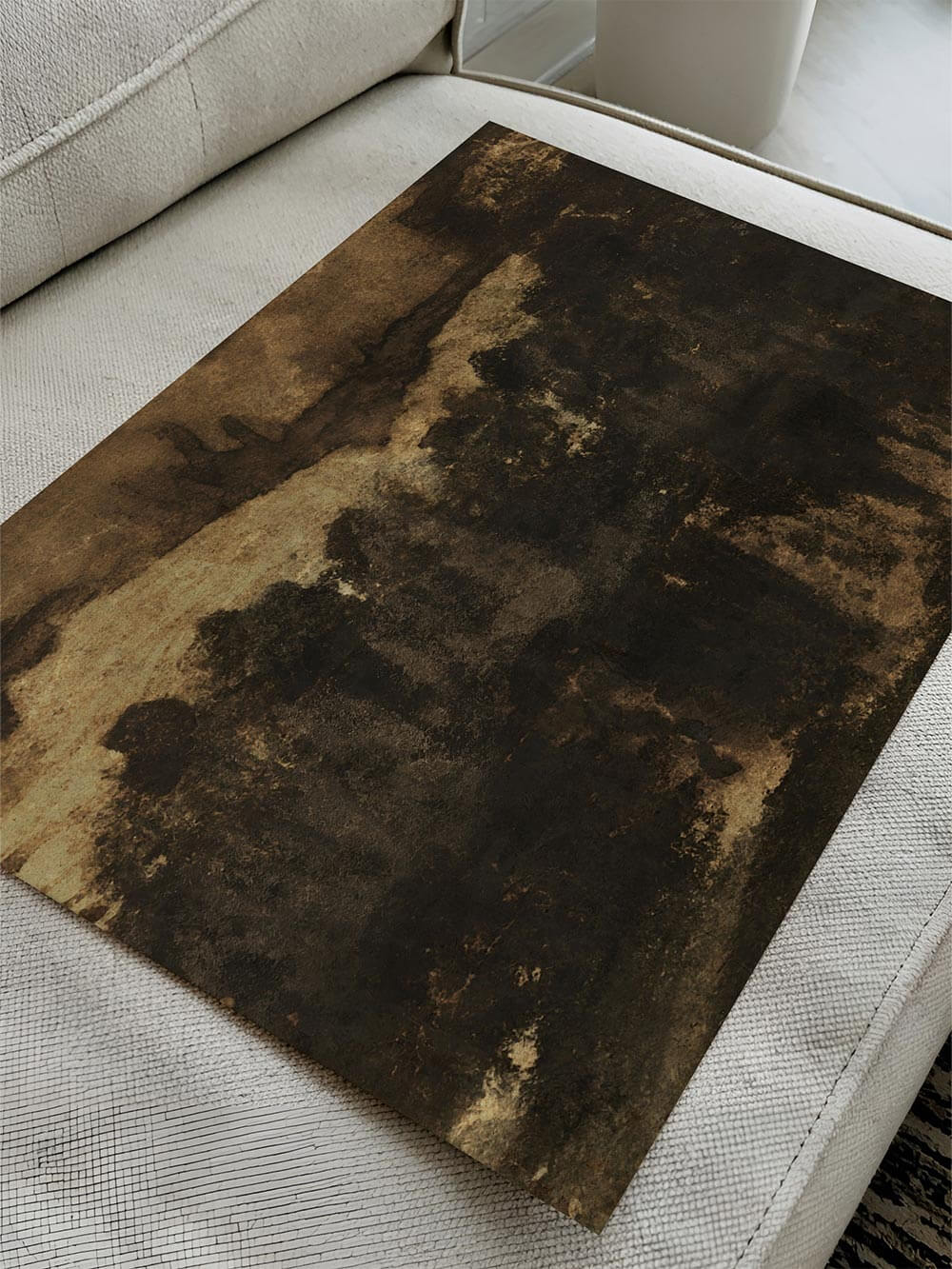Dunkler abstrakter Kunstdruck liegt auf einem hellen Sofa und zeigt eine vielschichtige Oberfläche mit erdigen Brauntönen und tiefen Schatten. Die schräge Perspektive betont die malerische Textur und eignet sich zur Präsentation von Poster oder Wandbild.
