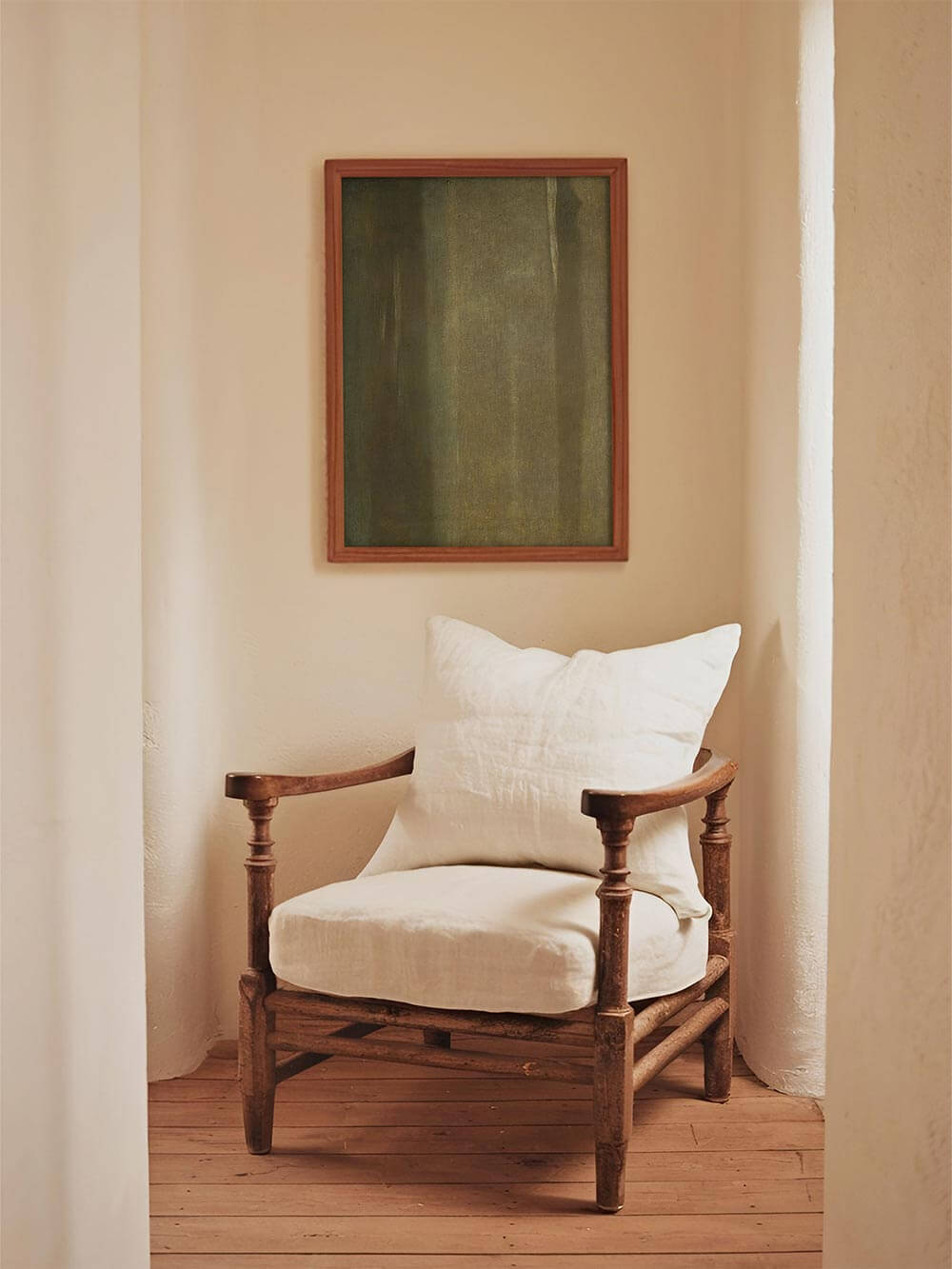 Abstrakter grüner Kunstdruck hängt in einer ruhigen Nische über einem Holzstuhl mit hellem Kissen und schafft mit seiner reduzierten Farbfläche eine warme, meditative Stimmung im Wabi-Sabi-Stil für Flur oder Leseecke.