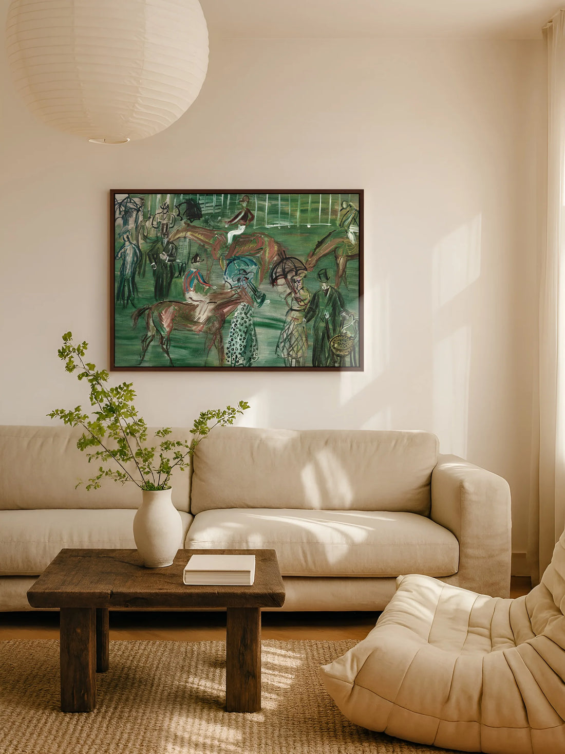 Das grüne Pferderennen Leinwandbild hängt im braunen Rahmen über einem beigen Leinensofa im Wohnzimmer. Davor steht ein rustikaler Couchtisch aus dunklem Holz mit einer weißen Vase und Zweigen, darüber hängt eine Papierlampe.