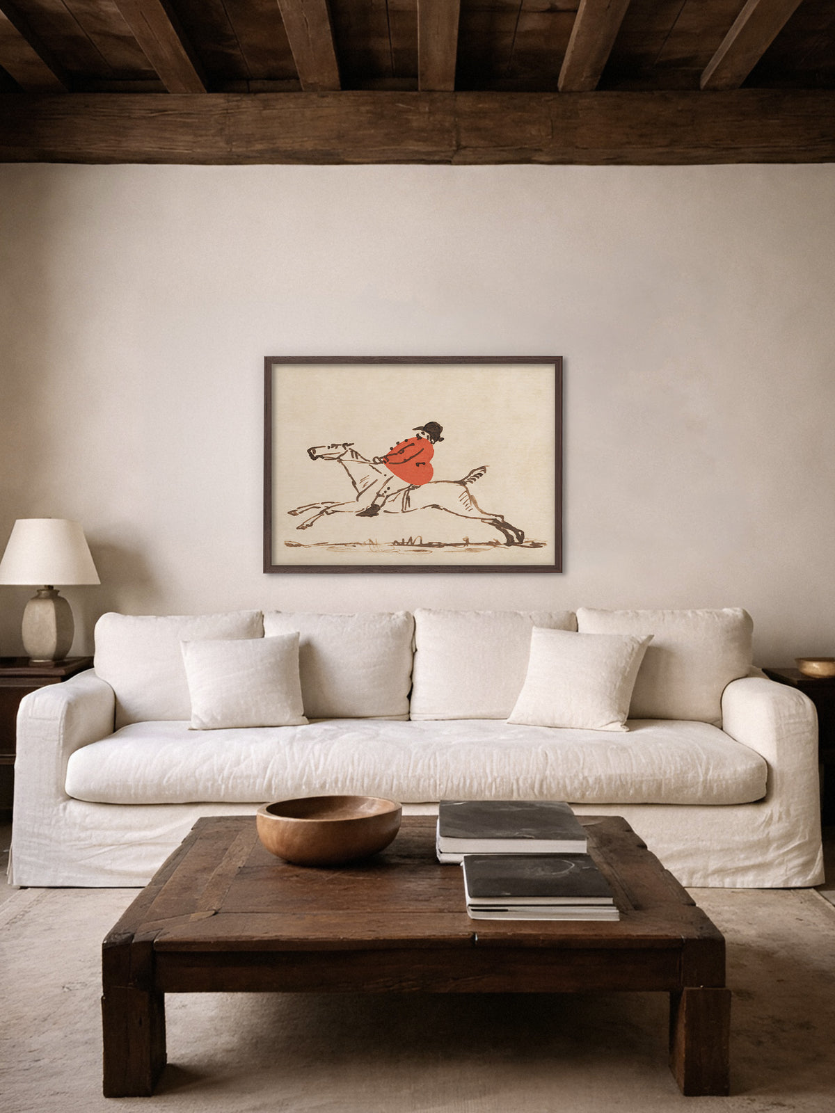 Reiter Poster im dunklen Holzrahmen über einem weißen Leinensofa in einem rustikalen Wohnzimmer mit dunklen Holzbalken, davor ein massiver Couchtisch mit Holzschale und Büchern. Die Vintage Zeichnung passt zu einer ruhigen Wabi Sabi Einrichtung.