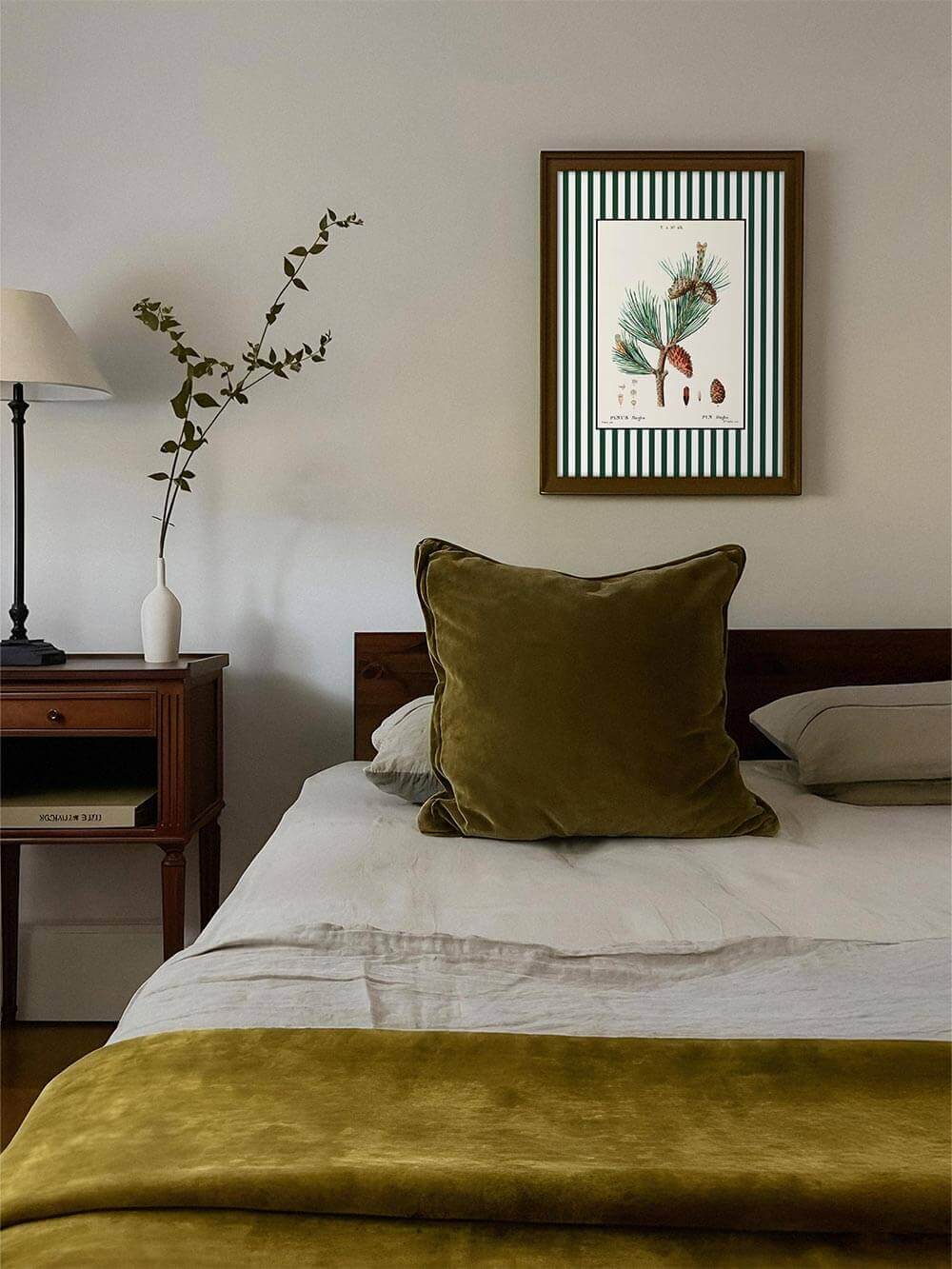 Botanischer Kunstdruck mit Kiefernzweig und Zapfen über einem Bett mit olivgrüner Decke und Kissen, ruhige Farbpalette, harmonisches Wandbild für natürliche Schlafzimmergestaltung.