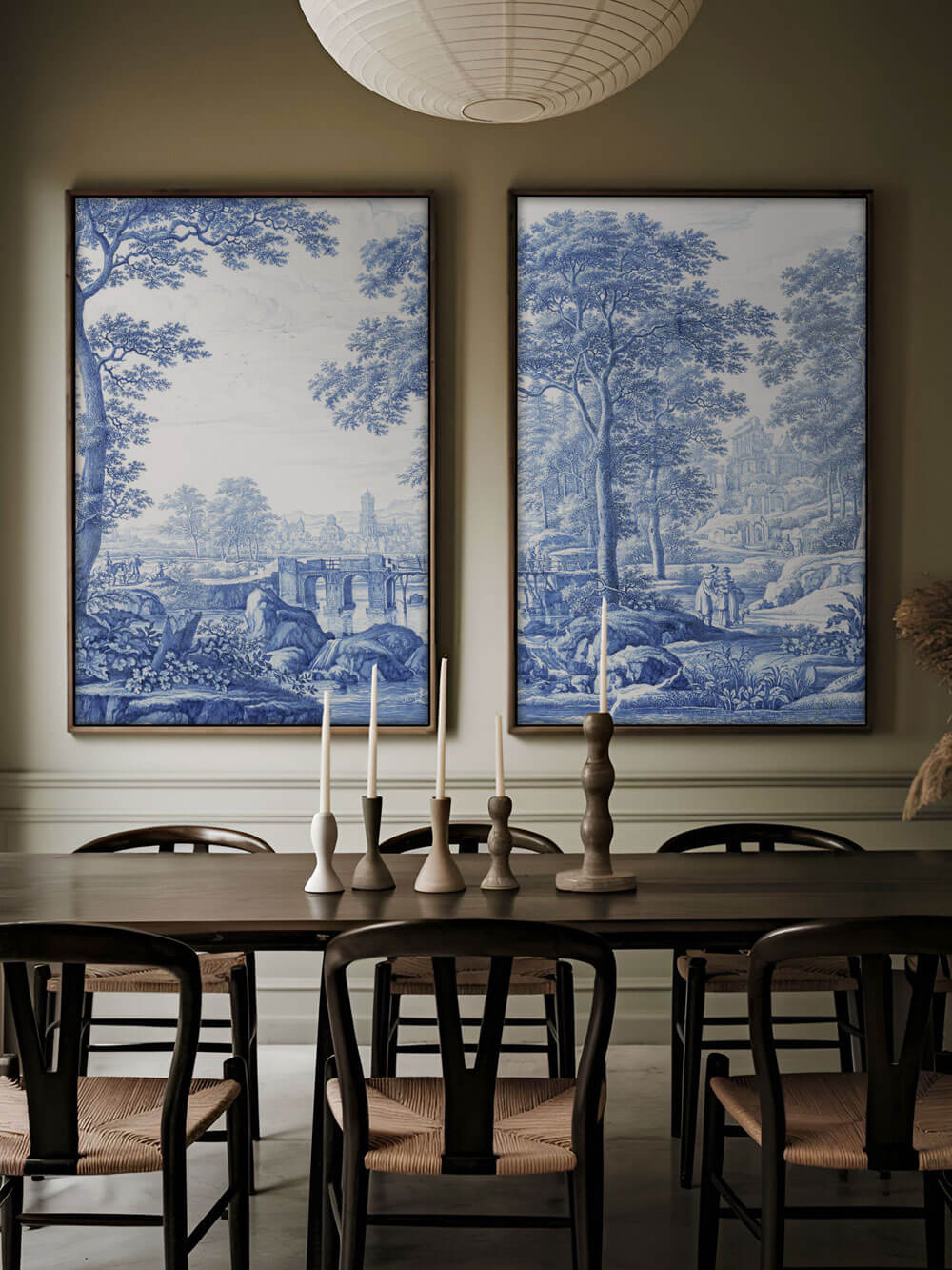 Zwei große, rahmengerahmte Kunstdrucke im Chinoiserie-Stil hängen nebeneinander über einem eleganten Esszimmer mit dunklem Holztisch und modernen Kerzenhaltern. Die Drucke zeigen detaillierte Landschaftsszenen in Blau-Weiß mit Bäumen, einer Brücke und Gebäuden. Die Kombination aus klassischem Kunstwerk und modernem Interieur schafft eine ruhige, stilvolle Atmosphäre.