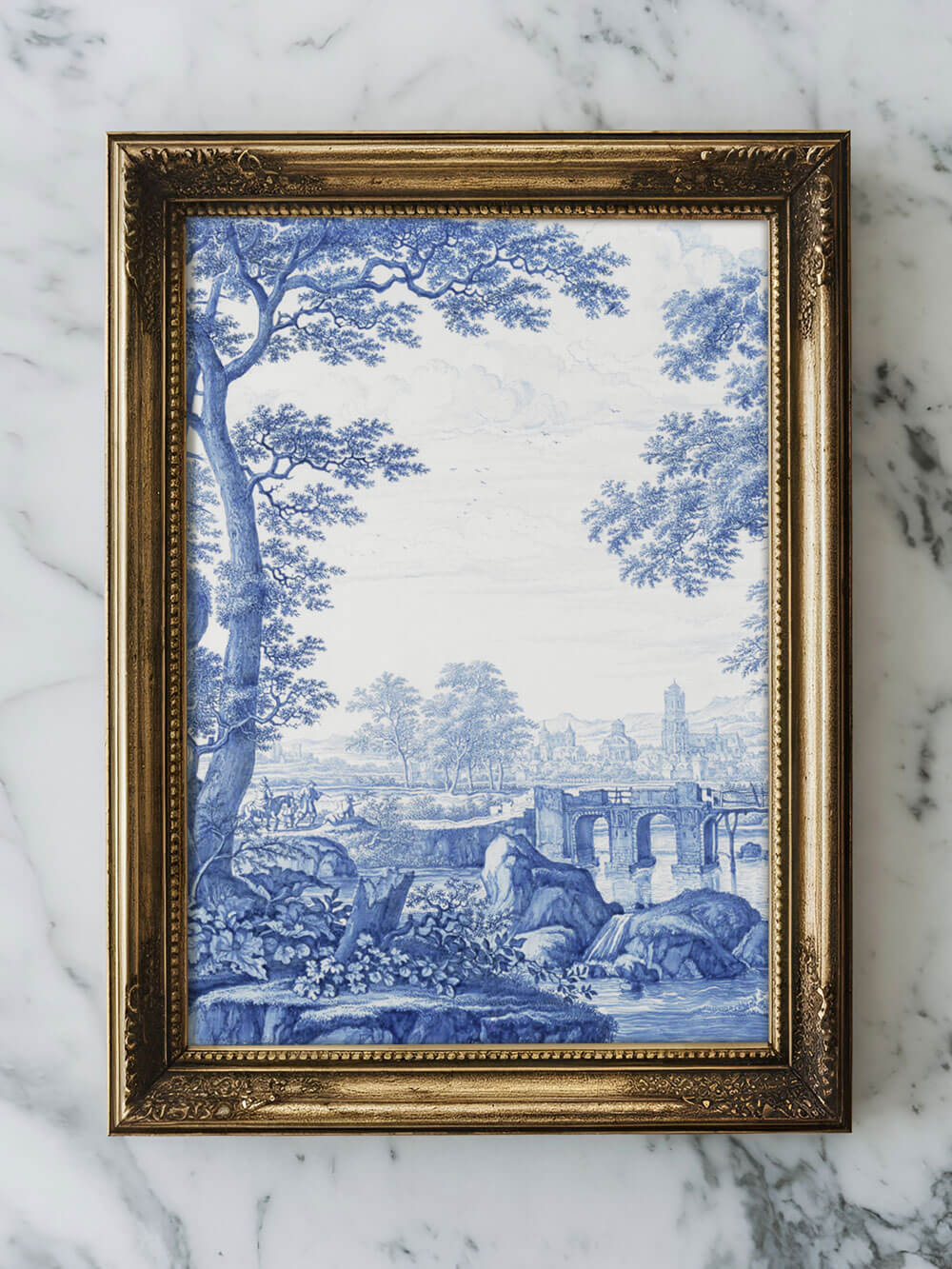 Nahaufnahme eines gerahmten Kunstdrucks einer Chinoiserie-Landschaft in Blau-Weiß. Der Rahmen ist aus goldenem, verzierten Holz und liegt auf einer Marmoroberfläche. Das Bild zeigt eine klassische, ruhige Landschaft mit Bäumen, Brücke und Fluss, die in weichen, blauen Farbtönen gestaltet ist.