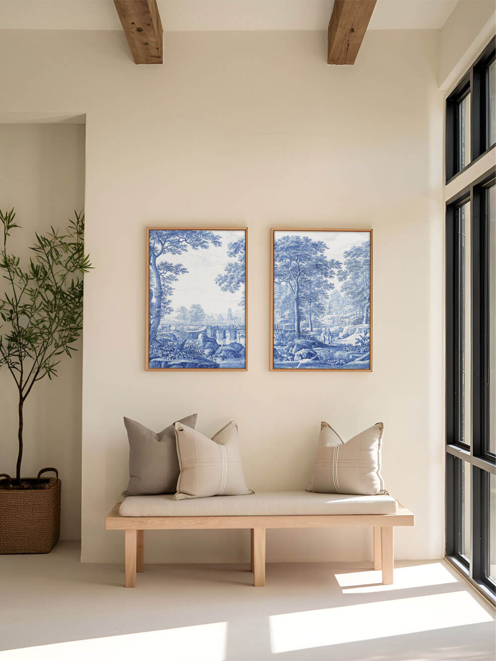Zwei blau-weiße Kunstdrucke im klassischen Stil hängen nebeneinander an einer beigen Wand in einem hellen Flur. Die detaillierten Landschaften zeigen Bäume, Brücken und ruhige Szenen aus der Natur. Eine schlichte Holzbank mit zwei beigefarbenen Kissen darunter verstärkt die ruhige, harmonische Stimmung.