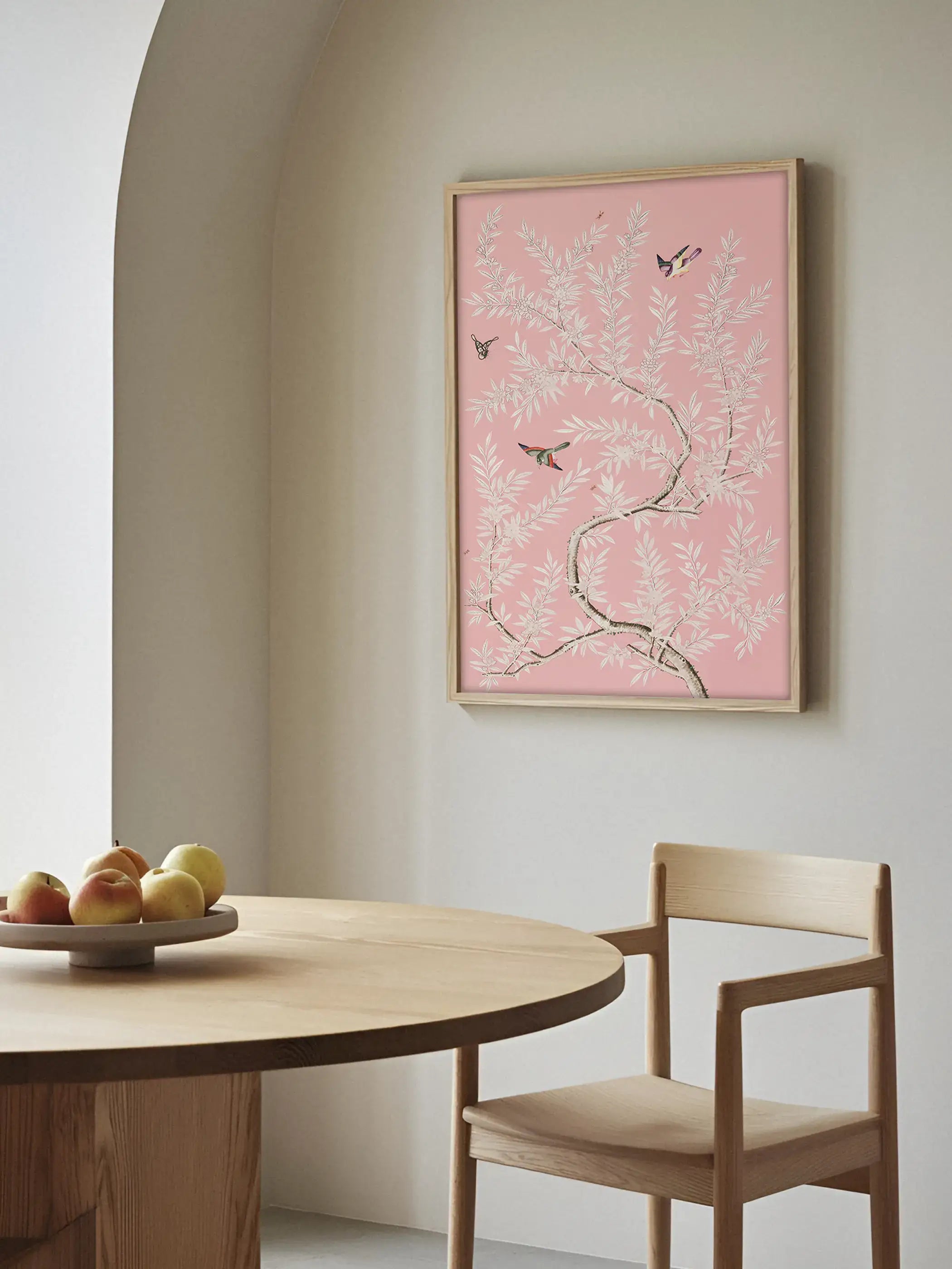 Ein rosa Chinoiserie Kunstdruck hängt über einem Holztisch in einem hellen Esszimmer mit natürlichen Materialien. Das botanische Wandbild setzt einen sanften Farbakzent und ergänzt moderne Wohnräume.