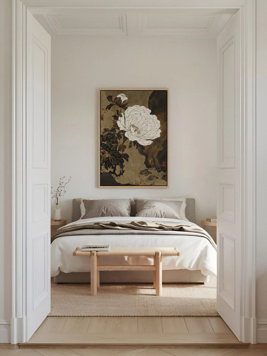 Gerahmtes Leinwandbild mit weißer Pfingstrose über einem Bett mit grauem Polsterkopfteil in einem hellen Altbauschlafzimmer mit Stuck und Holzbank. Das Japandi Wandbild setzt einen dunklen, ruhigen Akzent.