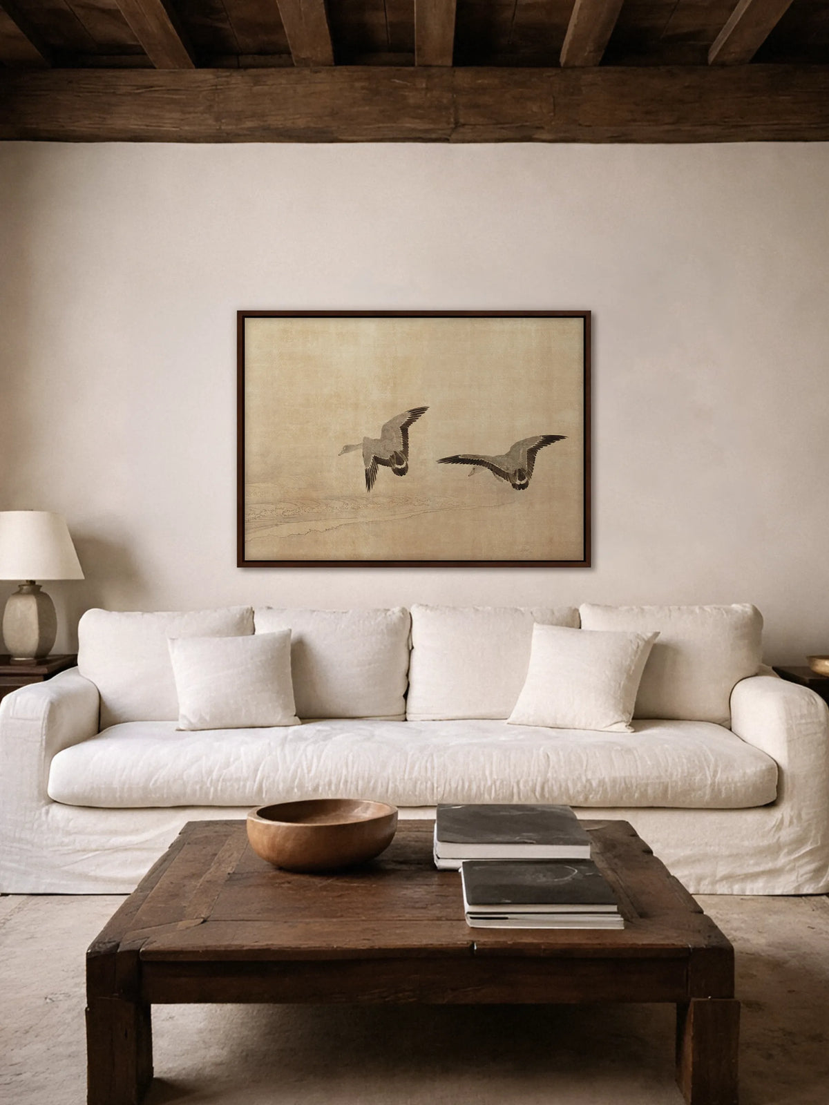 Das Wabi Sabi Leinwandbild mit zwei fliegenden Gänsen hängt im braunen Rahmen über einem weißen Leinensofa unter dunklen Holzbalken. Davor steht ein rustikaler Couchtisch aus altem Holz mit einer Holzschale und Büchern.