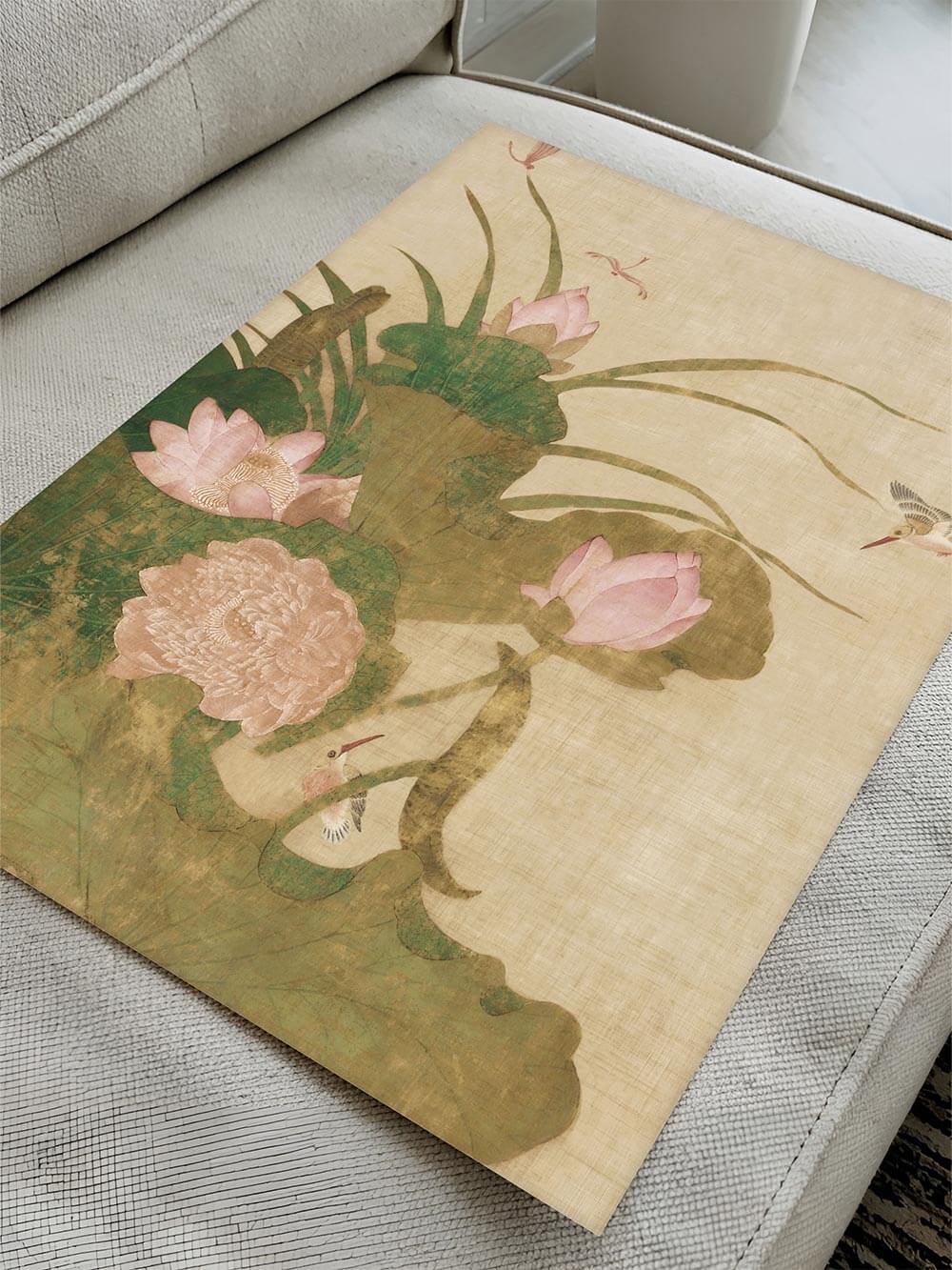 Chinoiserie Kunstdruck mit rosa Lotusblüten liegt auf einem hellen Sofa. Die detailreiche Illustration betont die feine Struktur und Farbnuancen des Motivs.