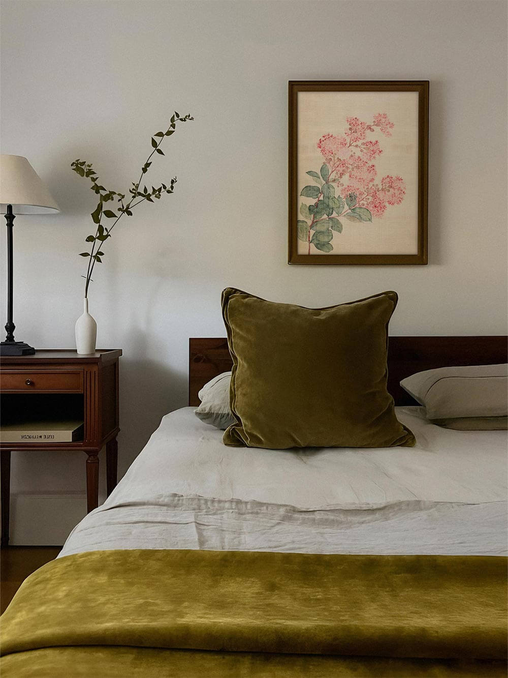 Ein floraler Kunstdruck mit rosa Blüten hängt gerahmt über einem Bett mit olivgrüner Decke und Kissen. Das Wandbild schafft eine ruhige, harmonische Atmosphäre.