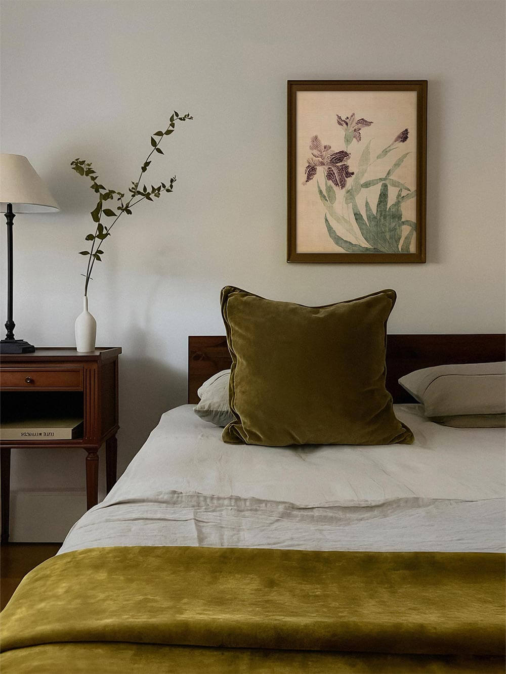 Der florale Kunstdruck mit violetten Irisblüten hängt gerahmt über einem Bett mit olivfarbenem Kissen. Die botanische Illustration verstärkt die harmonische Stimmung des Raumes.