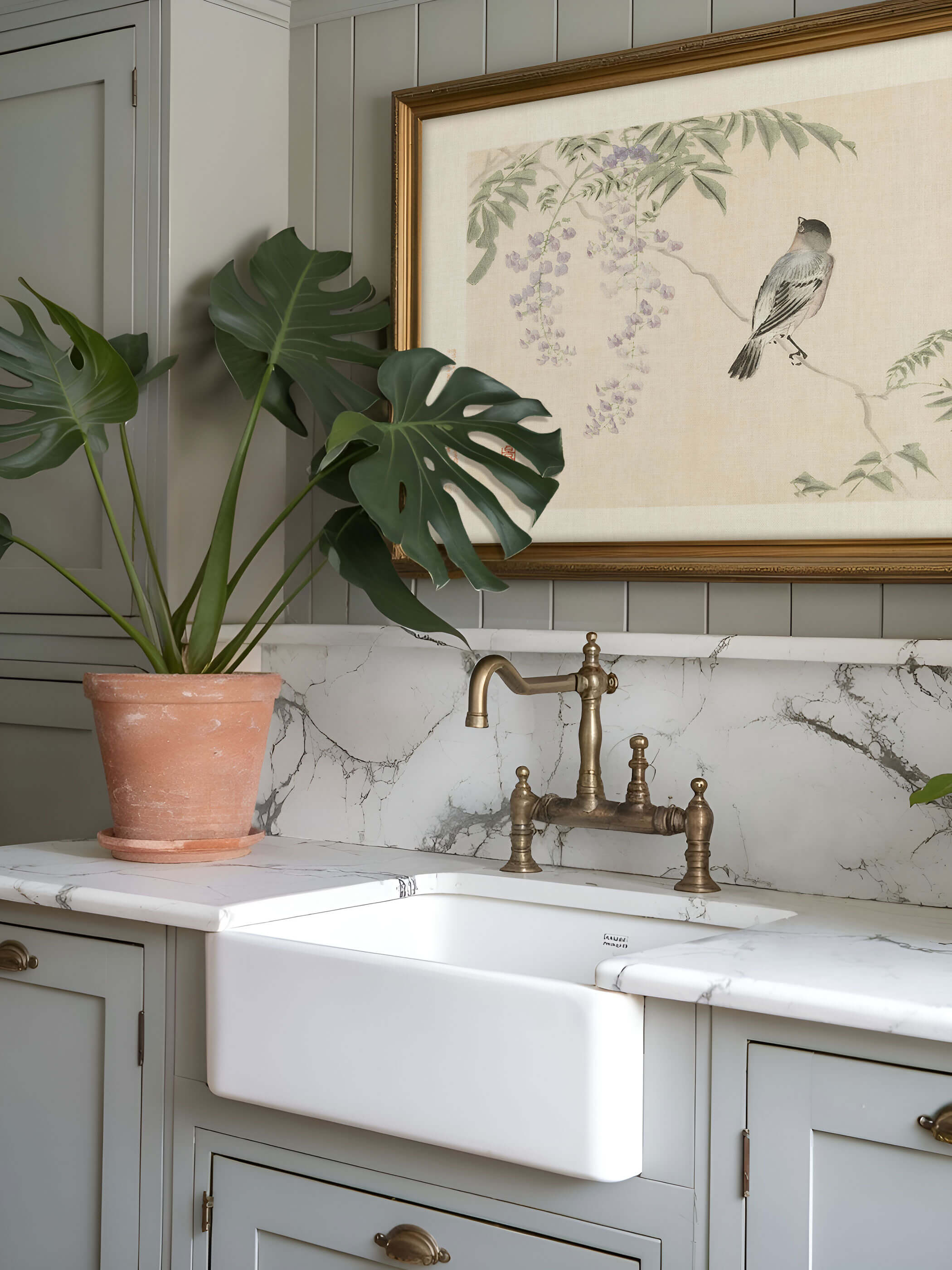 Gerahmter Chinoiserie Kunstdruck mit Vogel und Blauregen-Blüten hängt in schmalem Goldrahmen über einer Marmor-Küchenzeile mit Monstera und Messingarmatur. Das zarte Wandbild passt zu einem eleganten, naturnahen Küchenstil.
