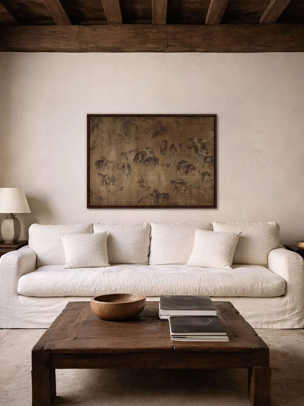 Das Wabi Sabi Leinwandbild mit Pferdeherde hängt im braunen Rahmen über einem weißen Leinensofa unter dunklen Holzbalken. Davor steht ein rustikaler Couchtisch aus altem Holz mit einer Holzschale und Büchern.