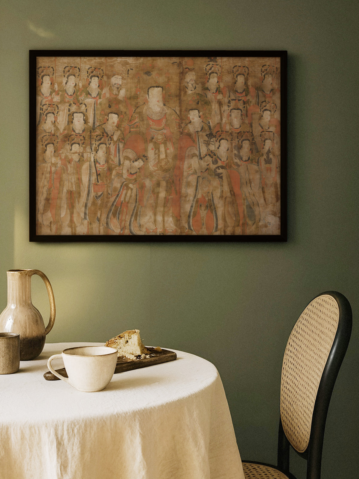 Chinoiserie Poster im schwarzen Rahmen an einer olivgrünen Wand über einem runden Tisch mit Leinentischdecke, Keramikkrug, Tasse und Kuchen, daneben ein Stuhl mit Wiener Geflecht. Das Rostrot der Figuren hebt sich warm vom moody Grün ab.