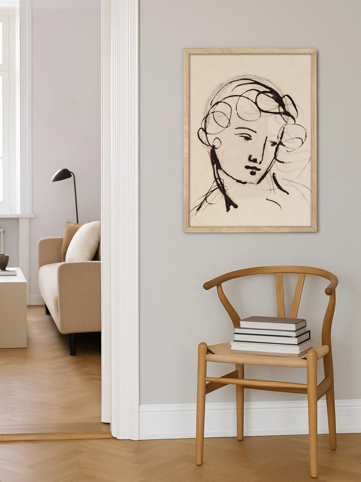 Frauenporträt als Poster im Rahmen aus hellem Holz über einem Holzstuhl mit geflochtener Sitzfläche und Bücherstapel, daneben der Blick in ein Wohnzimmer mit beigem Sofa. Die Vintage Zeichnung passt zum hellen Japandi Stil auf Fischgrätparkett.