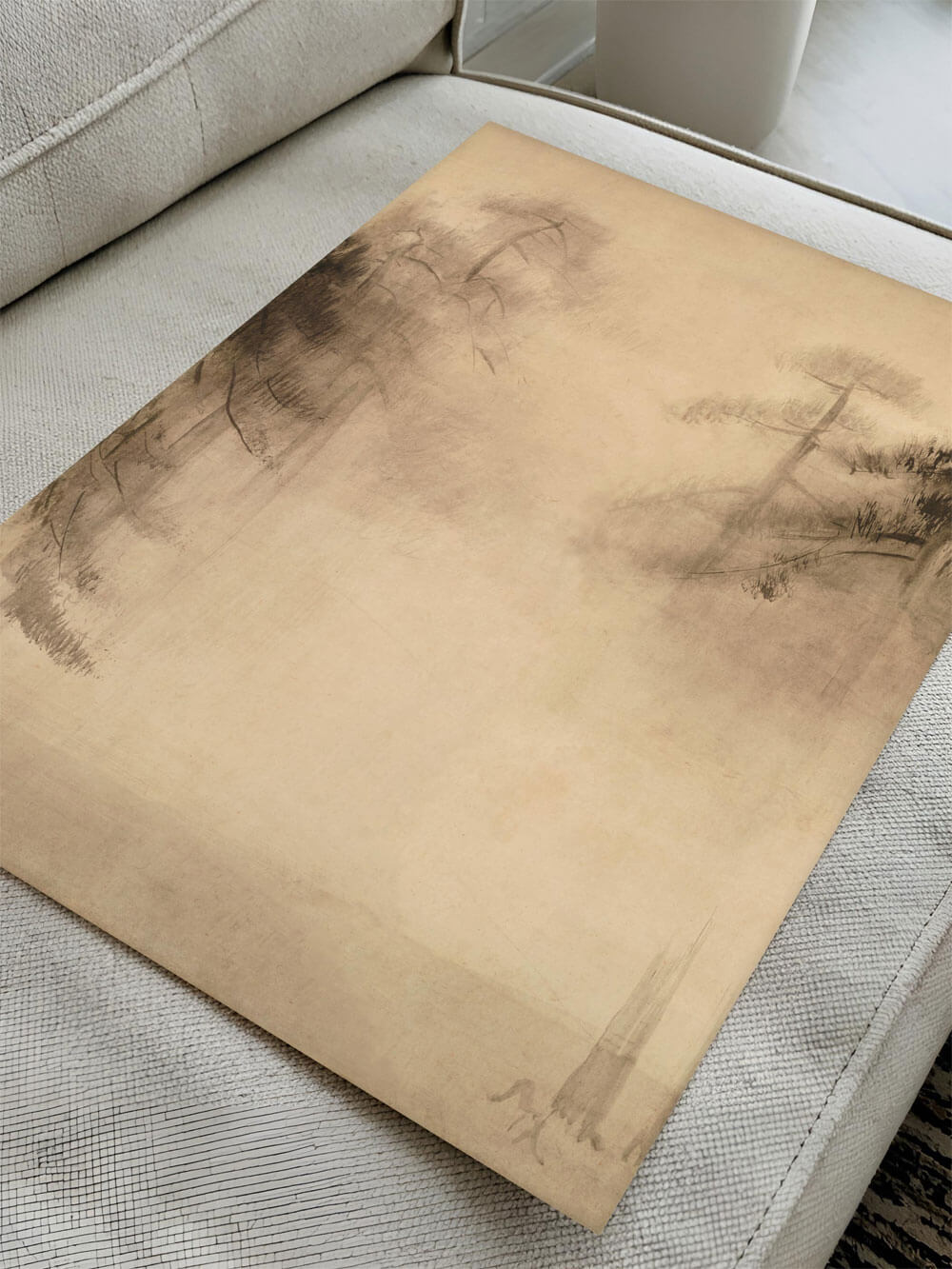 Eine Nahaufnahme eines Kunstposters im Wabi-Sabi-Stil, das auf einem hellen Sofa liegt. Das minimalistische Design zeigt zarte Kiefern und erzeugt eine ruhige, atmosphärische Stimmung.