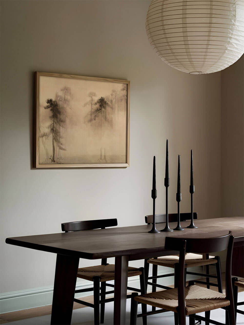 Ein minimalistischer Kunstdruck einer wabi-sabi-inspirierten Kiefernlandschaft hängt über einem modernen Esstisch mit dunklem Holz. Die neutrale Farbpalette des Raumes betont das ruhige, natürliche Design des Drucks.
