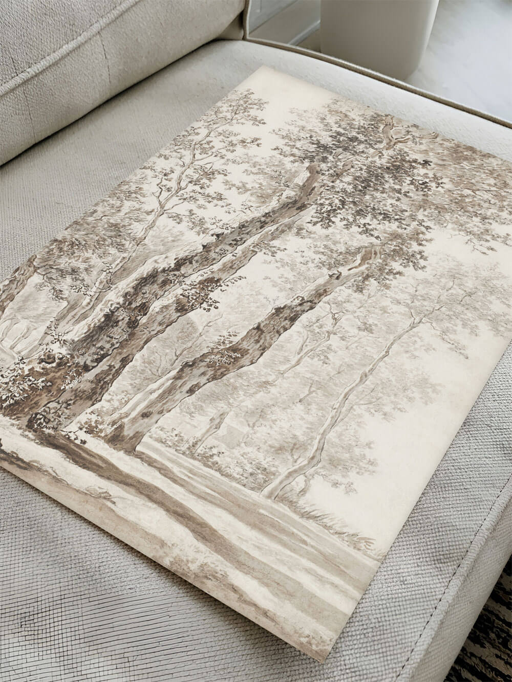 Detailaufnahme des Kunstdrucks einer antiken Baumzeichnung, liegend auf einem beigen Sofa. Die feinen Linien und sanften Schattierungen des Bildes sind sichtbar, ideal für Liebhaber klassischer Kunst.
