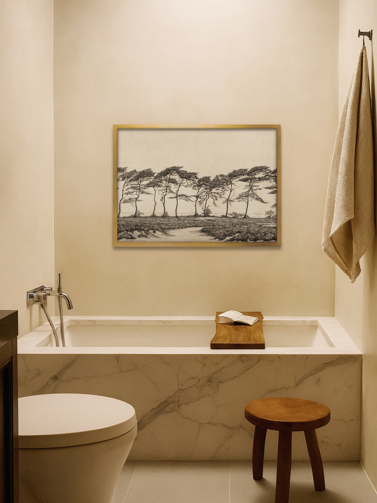 Kiefern Wandbild im goldenen Rahmen über einer Badewanne mit Marmorverkleidung, darauf ein Holzbrett mit offenem Buch, daneben ein Holzhocker und ein Leinenhandtuch. Die ruhige Landschaft passt zu einem warmen Badezimmer im Japandi Stil.