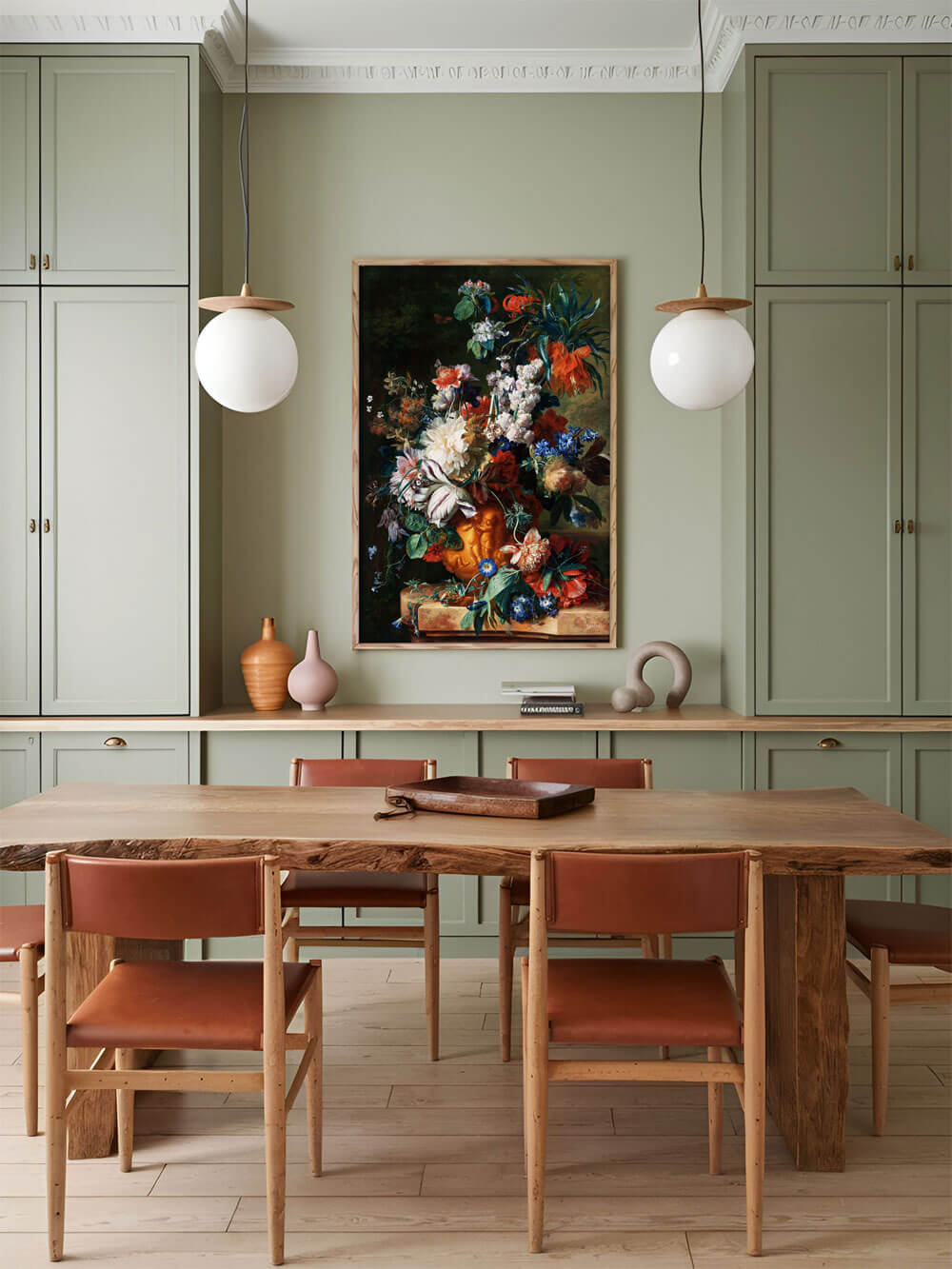 Ein barockes Blumenstillleben, eingerahmt in einem natürlichen Holzrahmen, hängt an einer grünen Wand über einem Esstisch mit Ledersesseln. Die kräftigen Blumenfarben – Rot, Blau, Weiß und Orange – kontrastieren mit dem dunklen Hintergrund und setzen lebendige Akzente in der schlichten Umgebung.