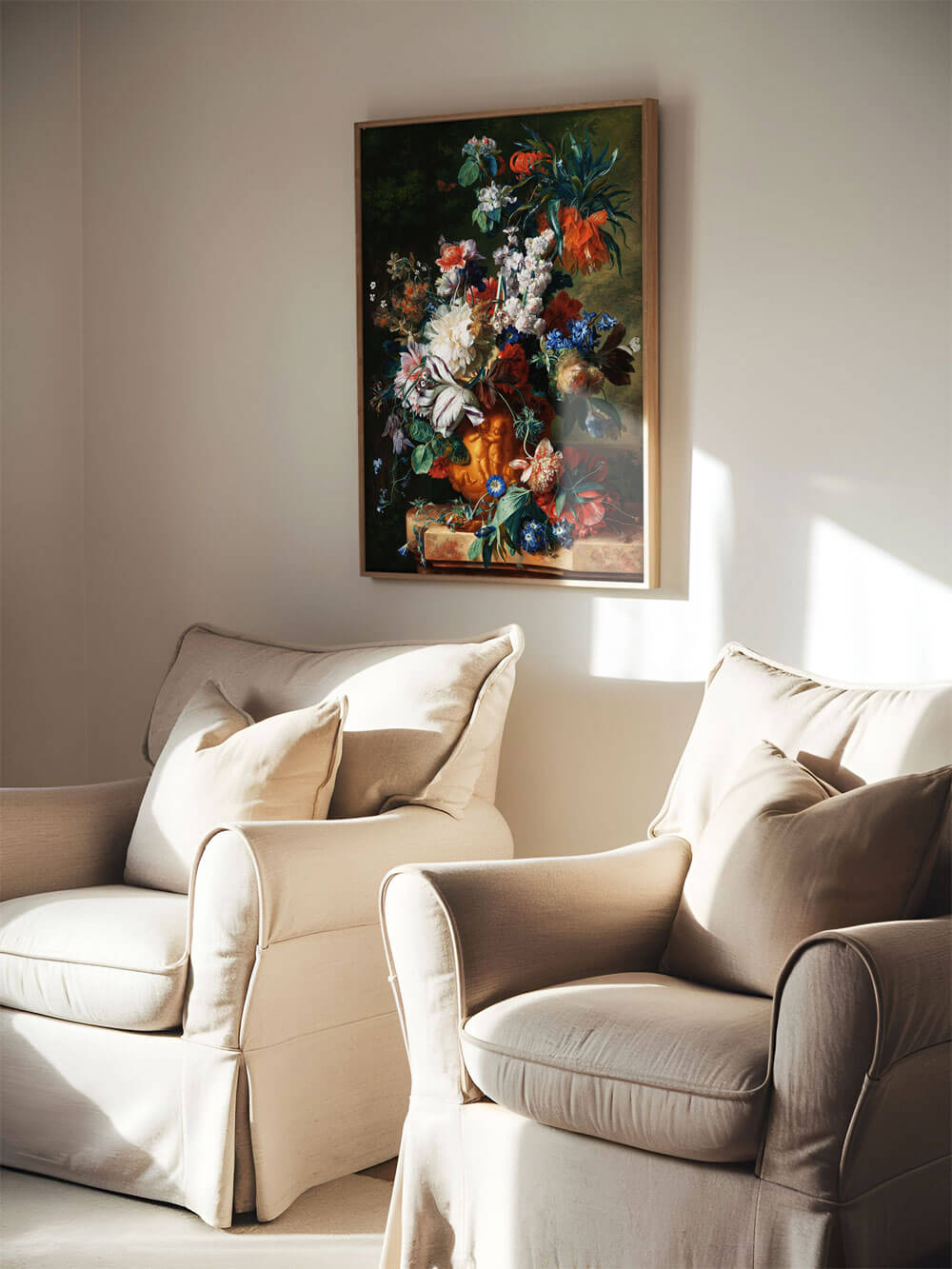 Ein barockes Blumenstillleben im Holzrahmen, das an einer hellen Wohnzimmerwand über zwei beigen Sesseln hängt. Der Kunstdruck zeigt eine opulente Blumenvase mit kräftigen Farben, darunter Rot-, Blau- und Weißtöne, die sich dramatisch vom dunklen Hintergrund abheben und eine klassische, elegante Stimmung schaffen.