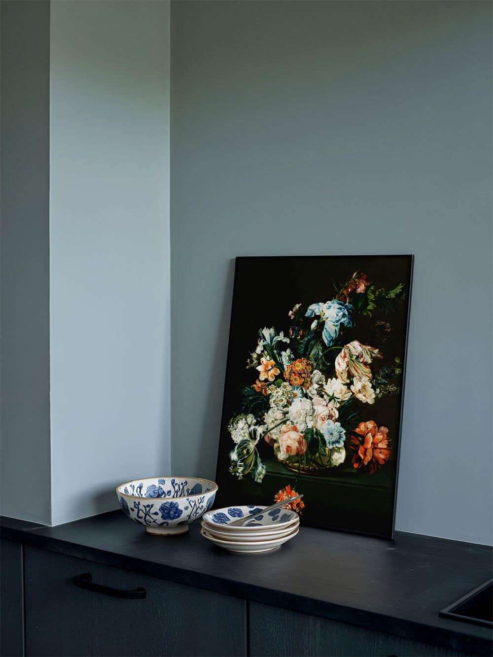 Ein Kunstdruck eines Barock-Blumenstilllebens steht leicht schräg an eine dunkelgraue Küchenwand gelehnt. Der Druck zeigt eine üppige Auswahl an Blumen in leuchtenden Farben, darunter Orange, Weiß, Rosa und Blau, auf schwarzem Hintergrund. Neben dem Bild sind stapelweise Teller und eine blau-weiß gemusterte Schale auf der dunklen Arbeitsplatte zu sehen.