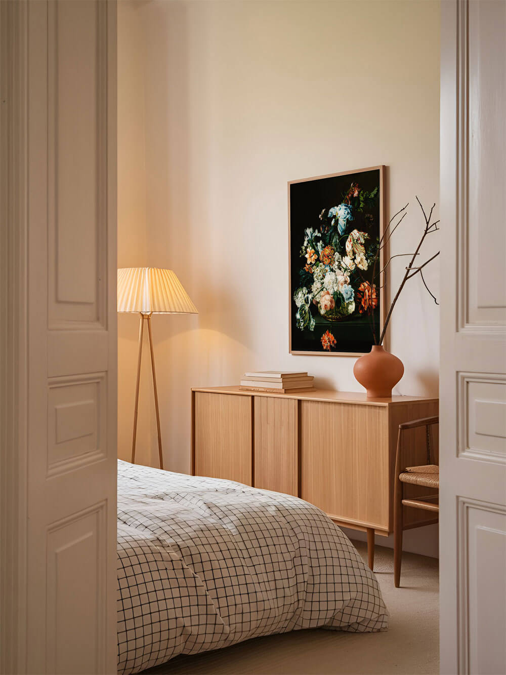 Ein Barock-Blumenstillleben hängt an einer beigen Wand in einem minimalistisch eingerichteten Schlafzimmer. Der Kunstdruck zeigt bunte Blumen auf schwarzem Hintergrund, darunter Rosen, Lilien und Pfingstrosen, in verschiedenen Farbtönen wie Rosa, Weiß und Blau. Darunter steht ein minimalistisches Sideboard aus hellem Holz, das mit einem Terrakotta-Vasenarrangement dekoriert ist, was dem Raum eine beruhigende, warme Atmosphäre verleiht.