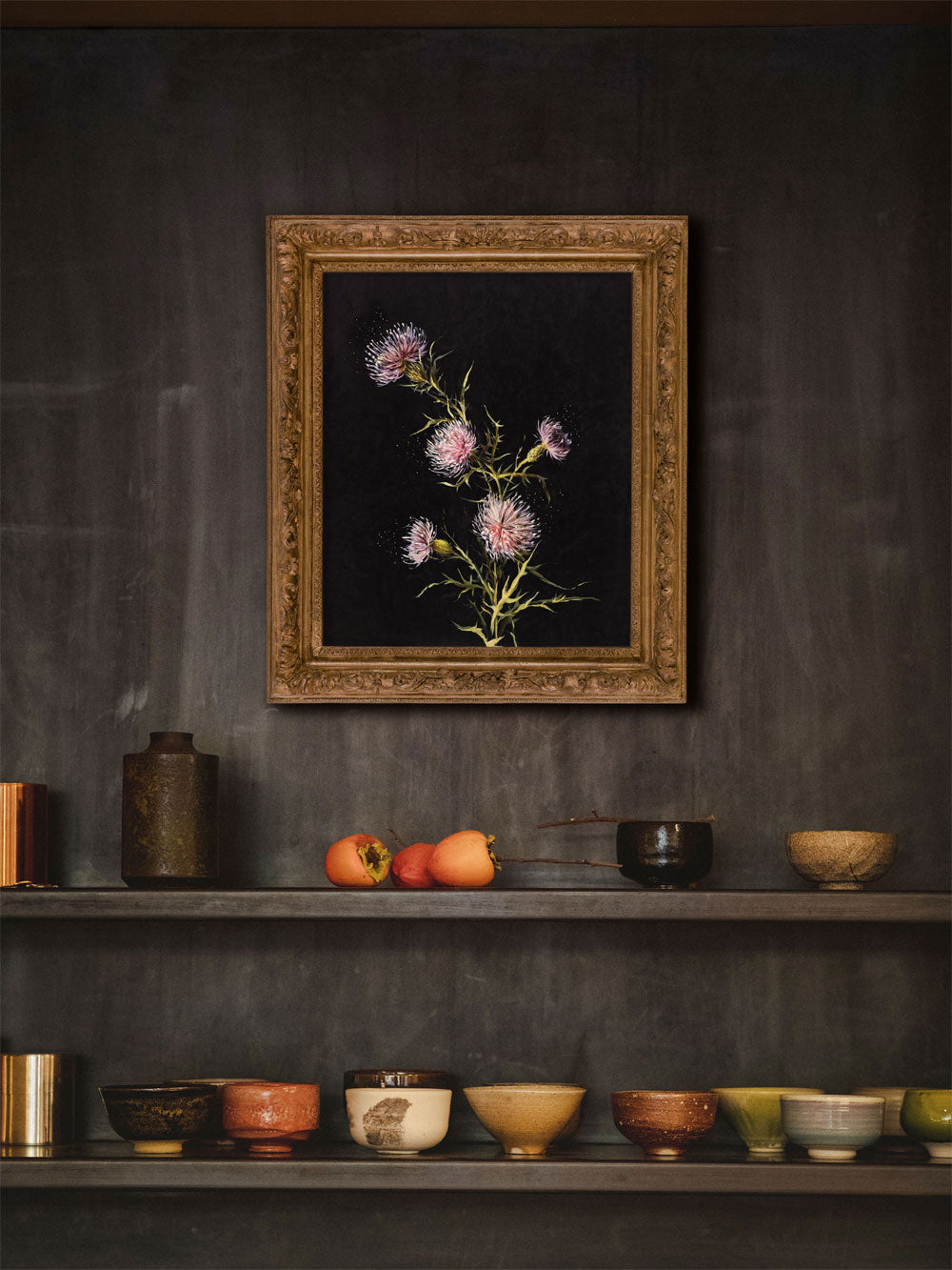 Distel-Poster im Vintage-Stil in einem goldverzierten Rahmen gefasst. Die zarten, stacheligen Blüten heben sich vor dem dunklen Hintergrund ab. Das Bild hängt an einer dunklen Wand, eingerahmt von Regalen mit kunstvoll arrangierten Keramikgefäßen, die dem Raum eine erdige, rustikale Eleganz verleihen.