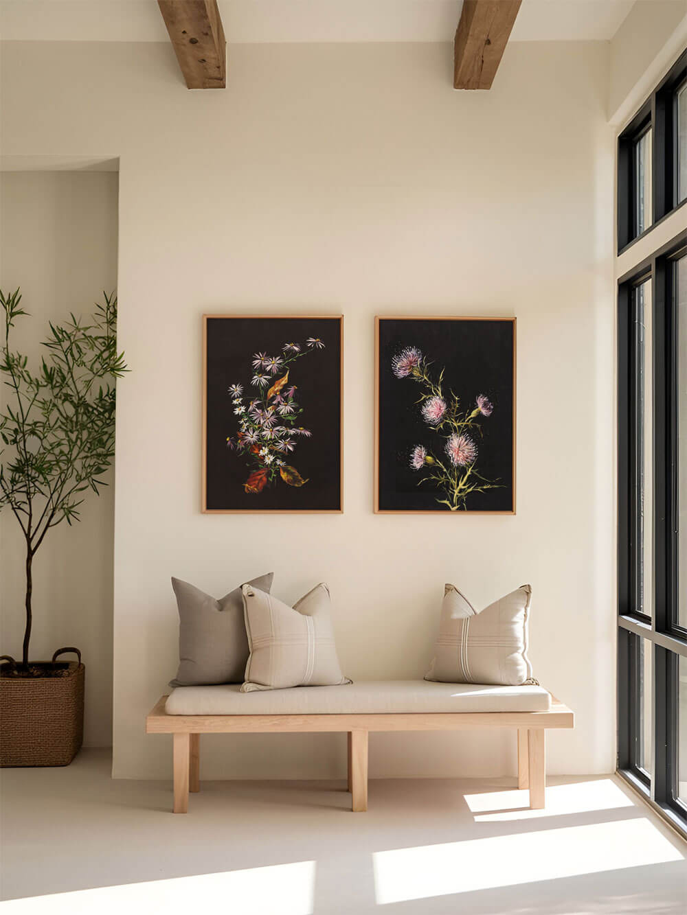 Zwei botanische Kunstdrucke nebeneinander, einer mit zarten pinken Distelblüten und der andere mit pastellfarbenen Blüten. Die Drucke hängen an einer cremefarbenen Wand über einer stilvollen, minimalistischen Holzbank, die mit beigen Kissen dekoriert ist. Natürliches Licht fällt durch große Fenster und betont die weichen Töne und Details der Drucke.